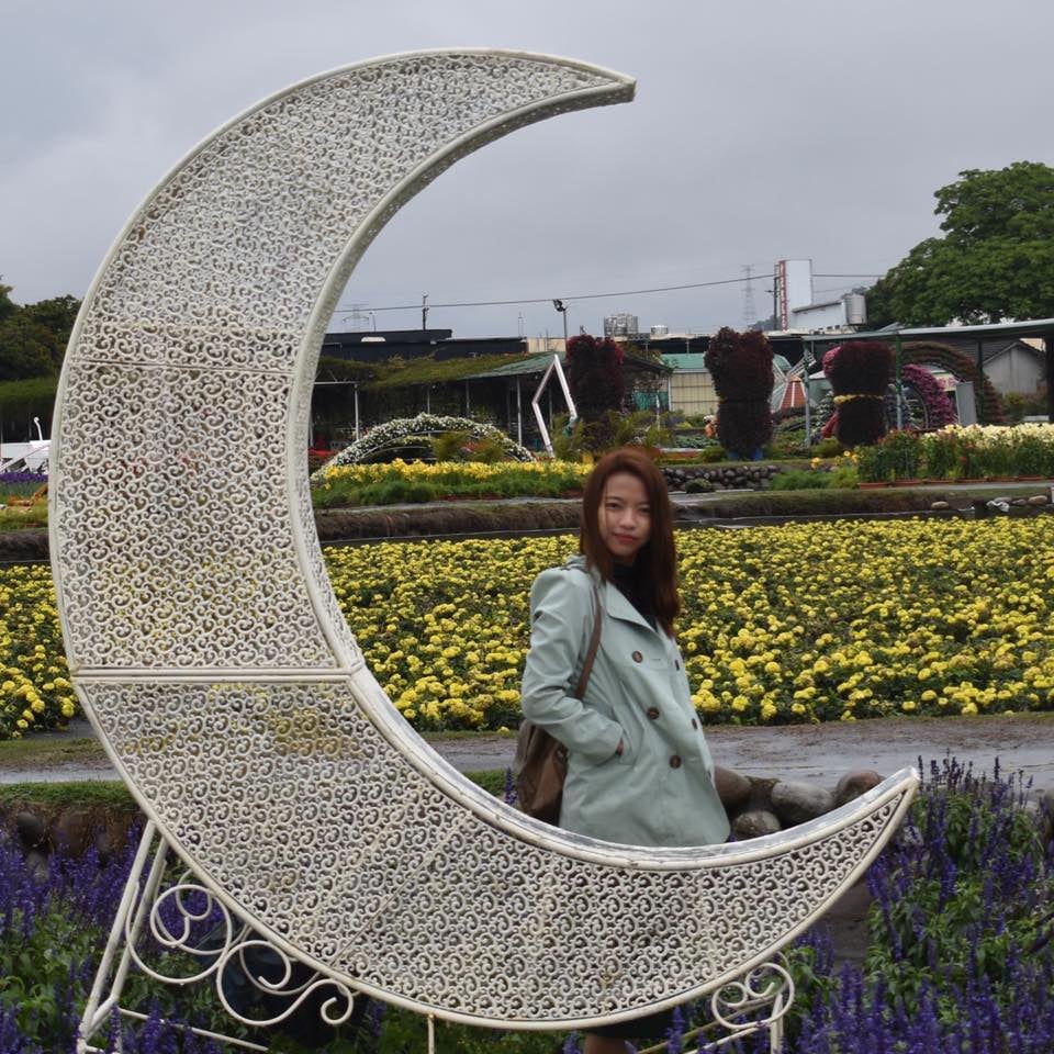 Woman in a light jacket stands near a decorative crescent moon in a garden with yellow flowers and overcast sky.