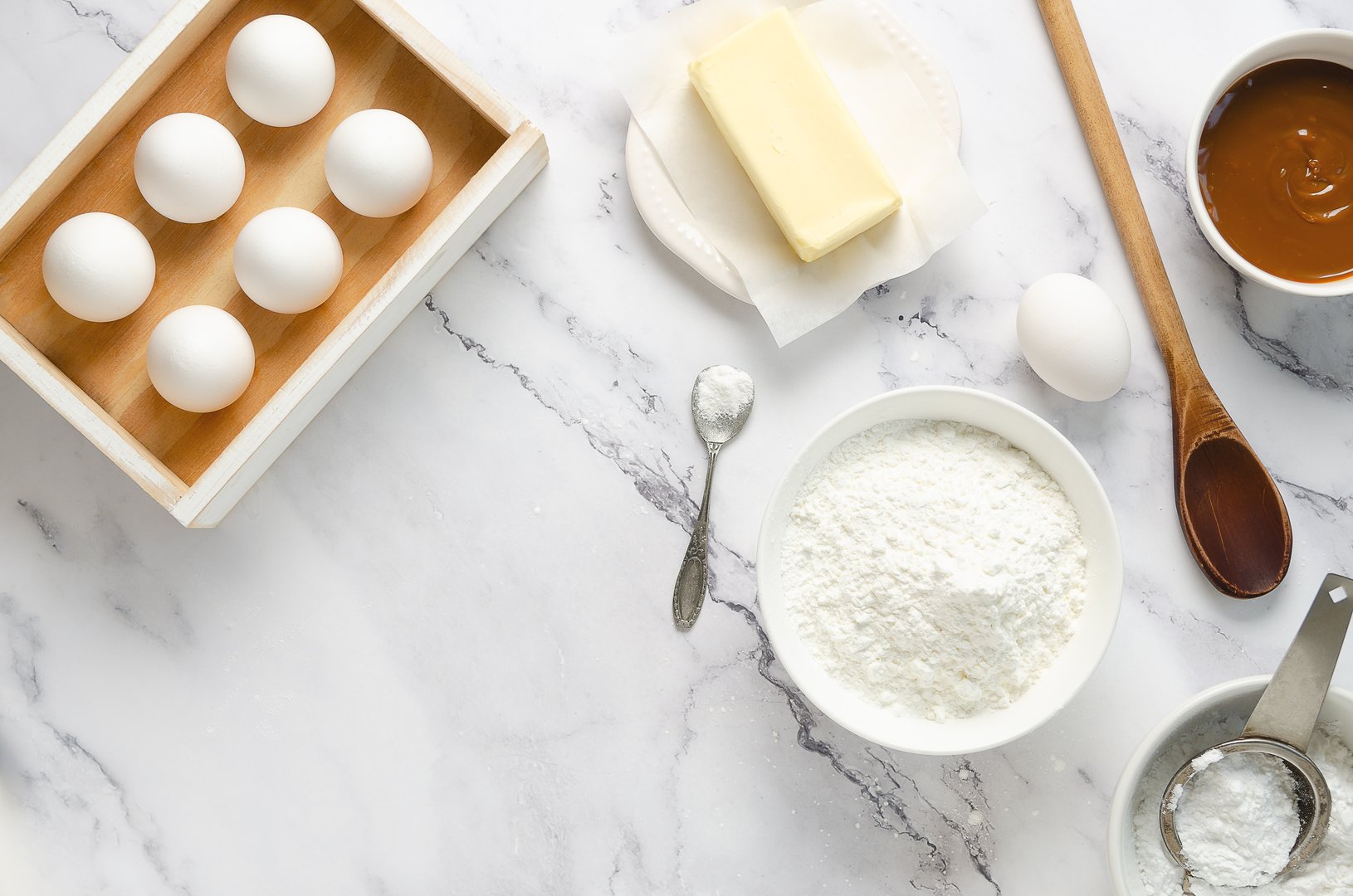 Preparing the ingredients for making cookies and corn starch alfajores. Eggs, butter, corn starch, powder sugar, baking powder, and dulce de leche on a white marble background.