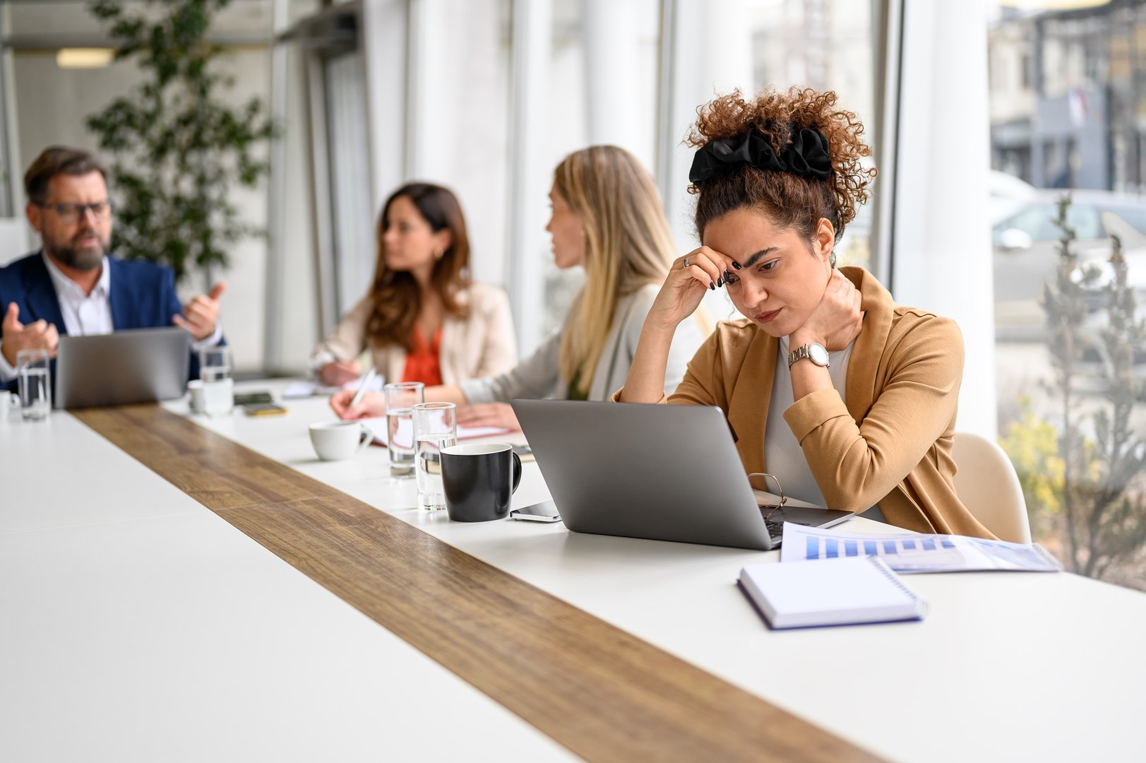 Stressed businesswoman focusing on her laptop while colleagues discussing and collaborating in the background during meeting