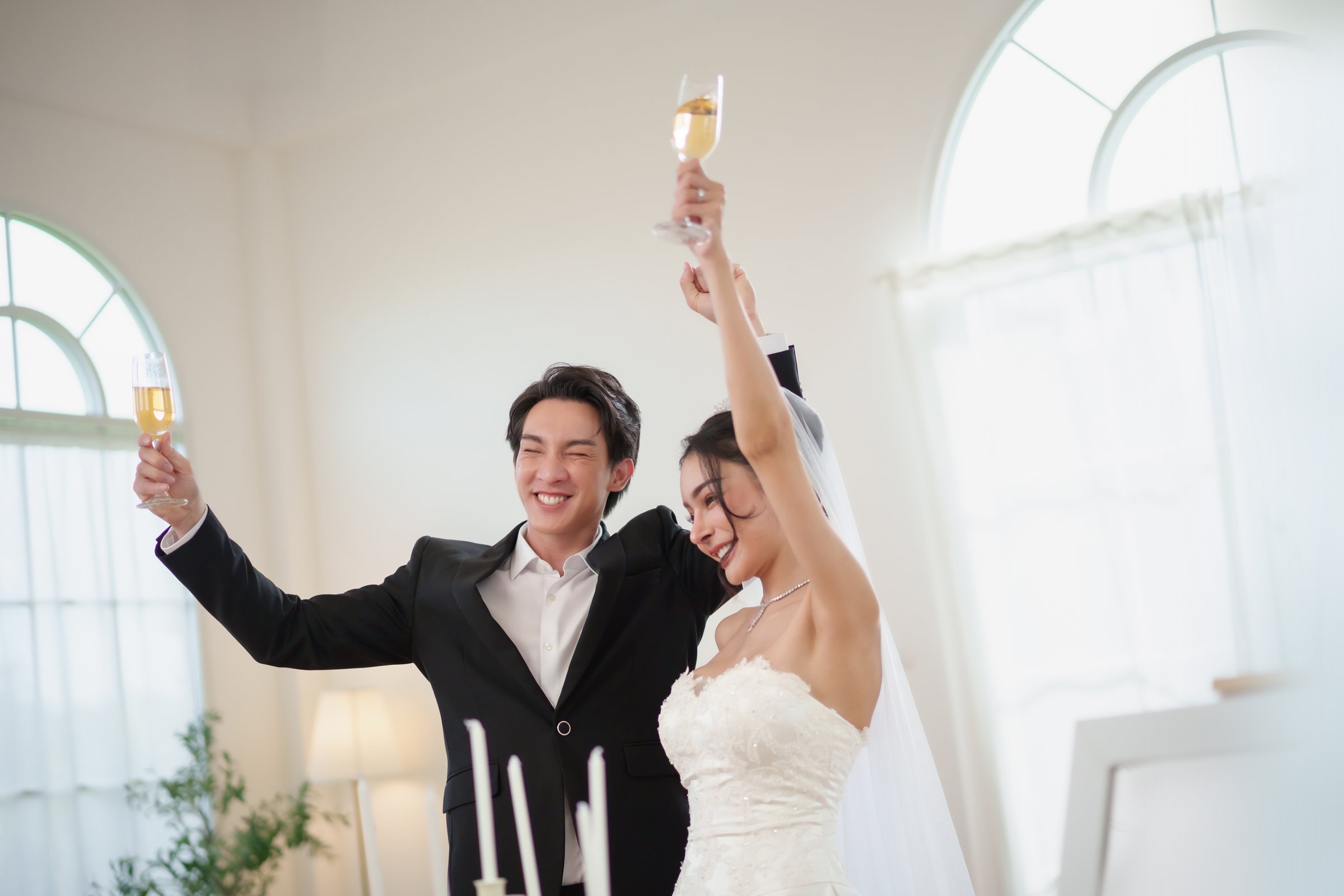 Happy Asian bride and groom celebrate wedding together with champagne toast inside studio, showing love and joy and raising glasses with happiness and cheers