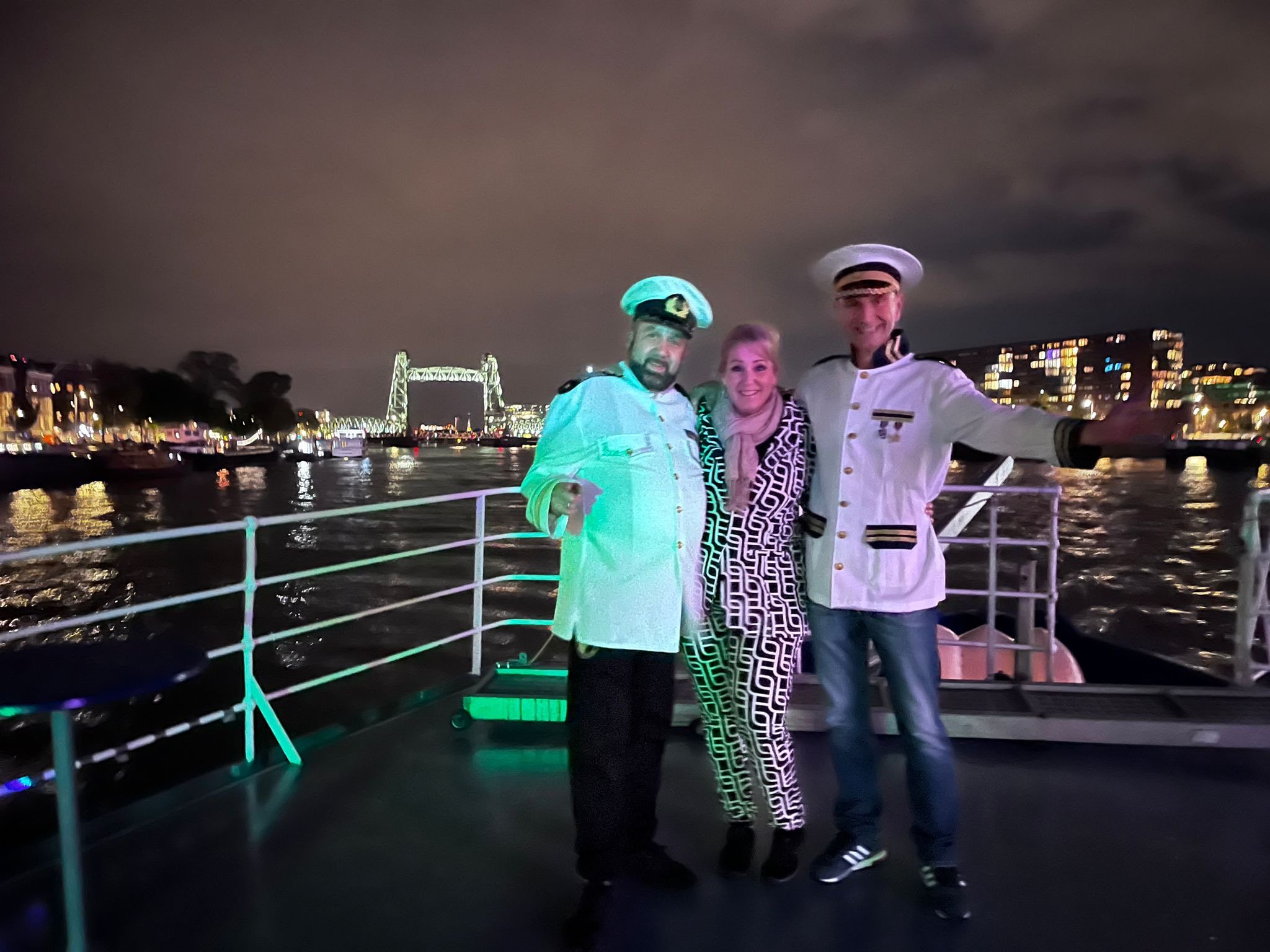 Three people in themed outfits posing on a boat at night, with a cityscape and bridge visible in the background.