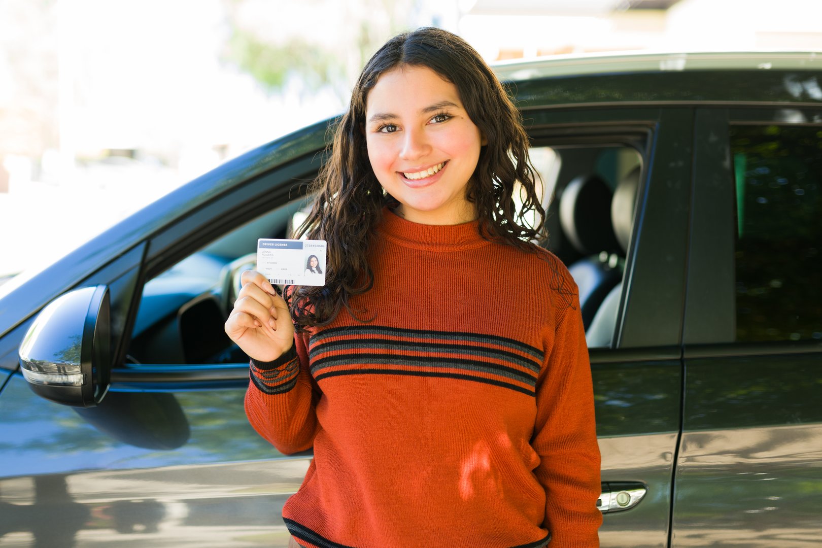 Cute and cheerful teenager holding her driving license while standing next to her new car outdoors