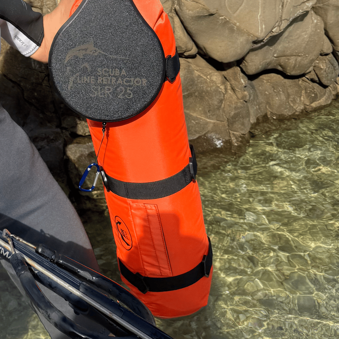 Person holding an orange scuba line retractor bag near rocky shore and clear water.
