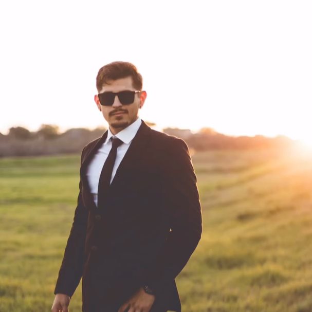 Man in sunglasses wearing a suit stands in a field at sunset, with a sunny sky and grassy landscape in the background.