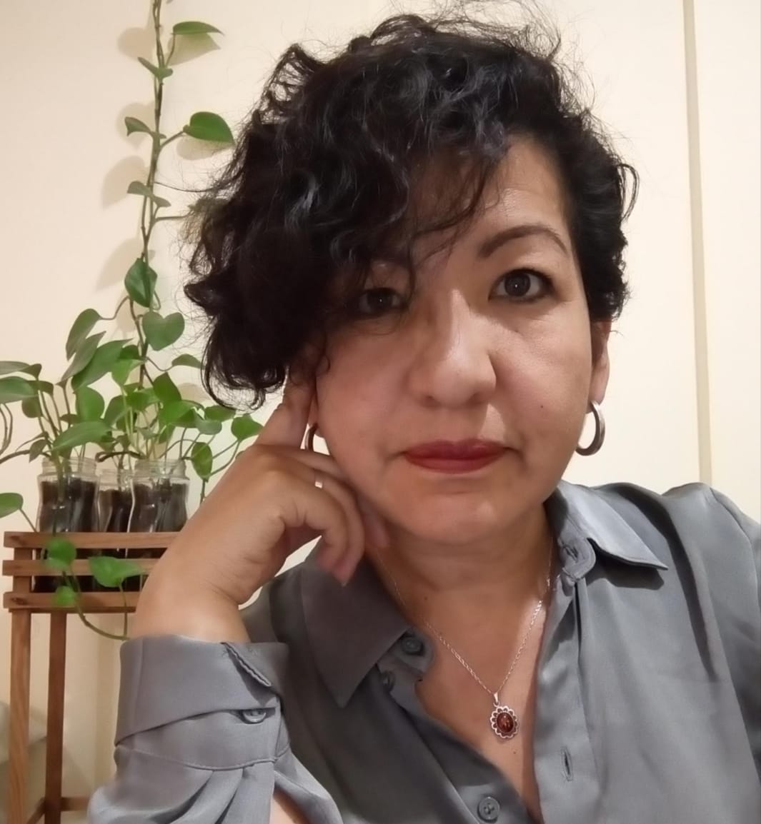 Person with short curly hair and silver hoop earrings poses thoughtfully in front of a plant, wearing a gray blouse.