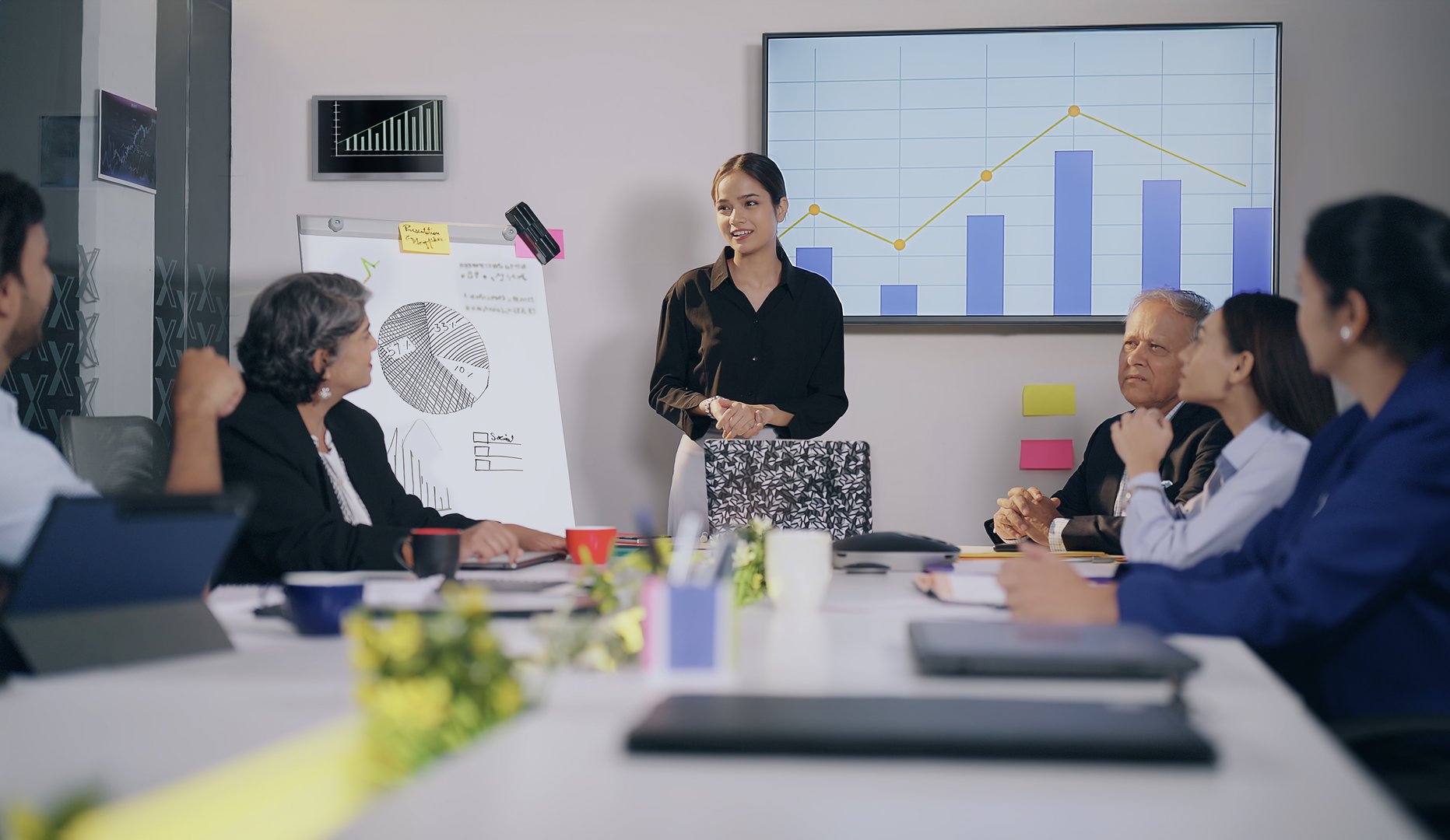 Indian business woman team leader show growth chart ideas whiteboard discuss market graph data at workplace. Young adult board of directors with expert staff worker lady group explaining presentation use TV screen boardroom office
