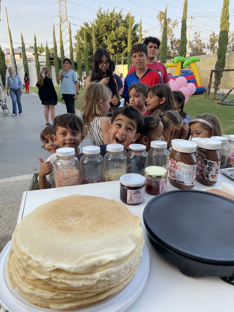 Children excitedly gather around a table with pancakes, jars of sprinkles, and toppings at an outdoor event.