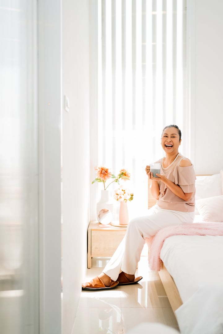 Happy middle-aged Asian woman enjoys a cup of organic soy milk in a bright, cozy bedroom. Embracing self-care, wellness, and a positive lifestyle. Perfect for concepts of healthy aging and relaxation.