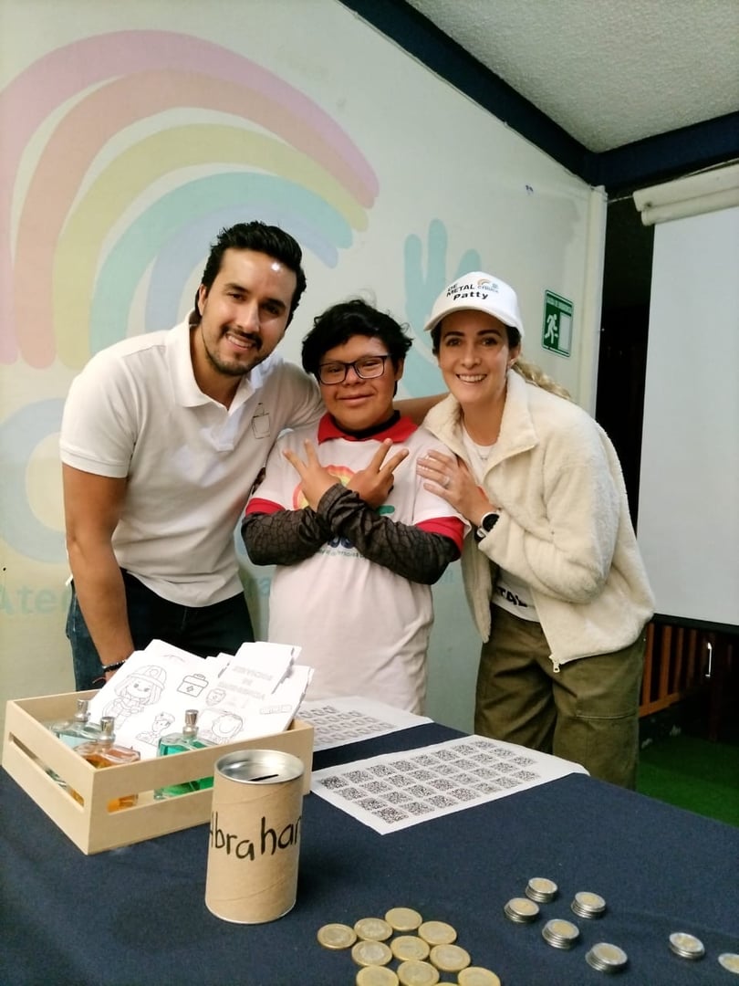 Three people smiling at a table with crafts and coins, standing indoor near a painted wall.