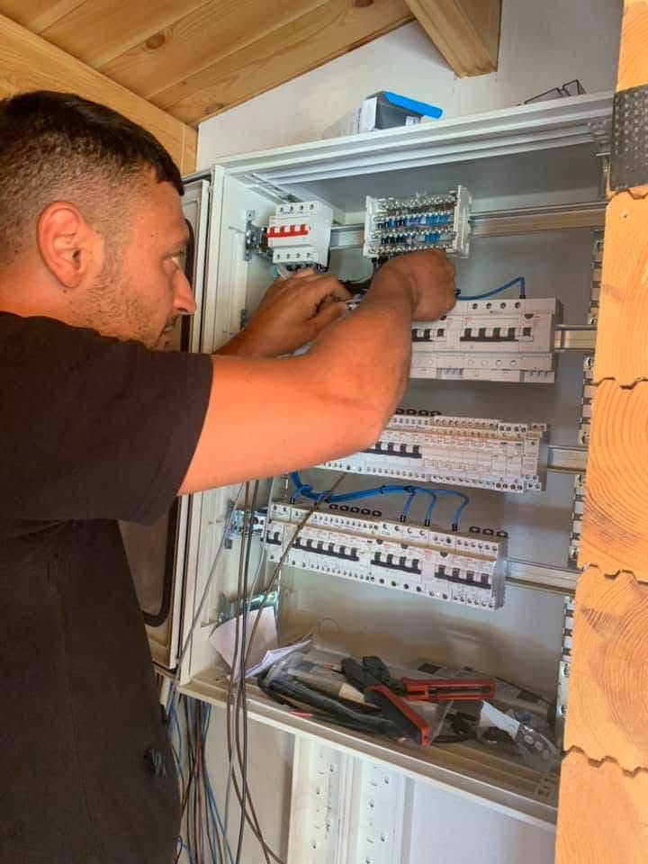 Electrician working on a circuit breaker panel, arranging wires with tools and paperwork nearby.