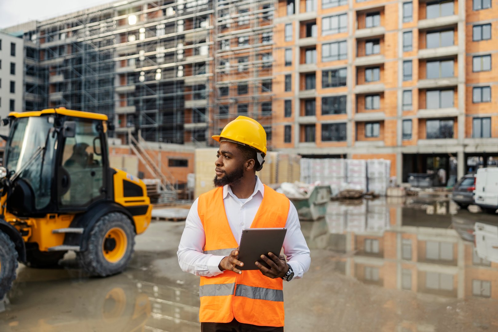 An african american project contractor in protective uniform standing at construction site with tablet in hands and monitoring works.