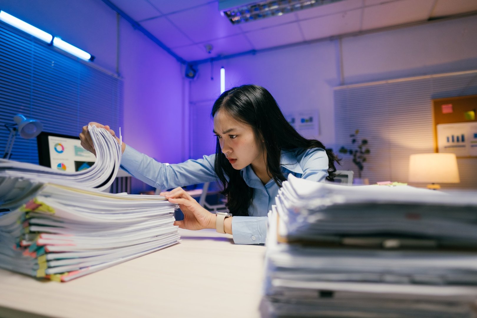Young asian businesswoman searching through stacks of paperwork at her desk late at night, struggling to find important documents in dimly lit office