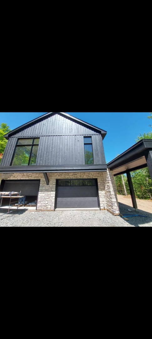 Modern two-story house with a black exterior, large windows, and a stone base, set against a bright blue sky.
