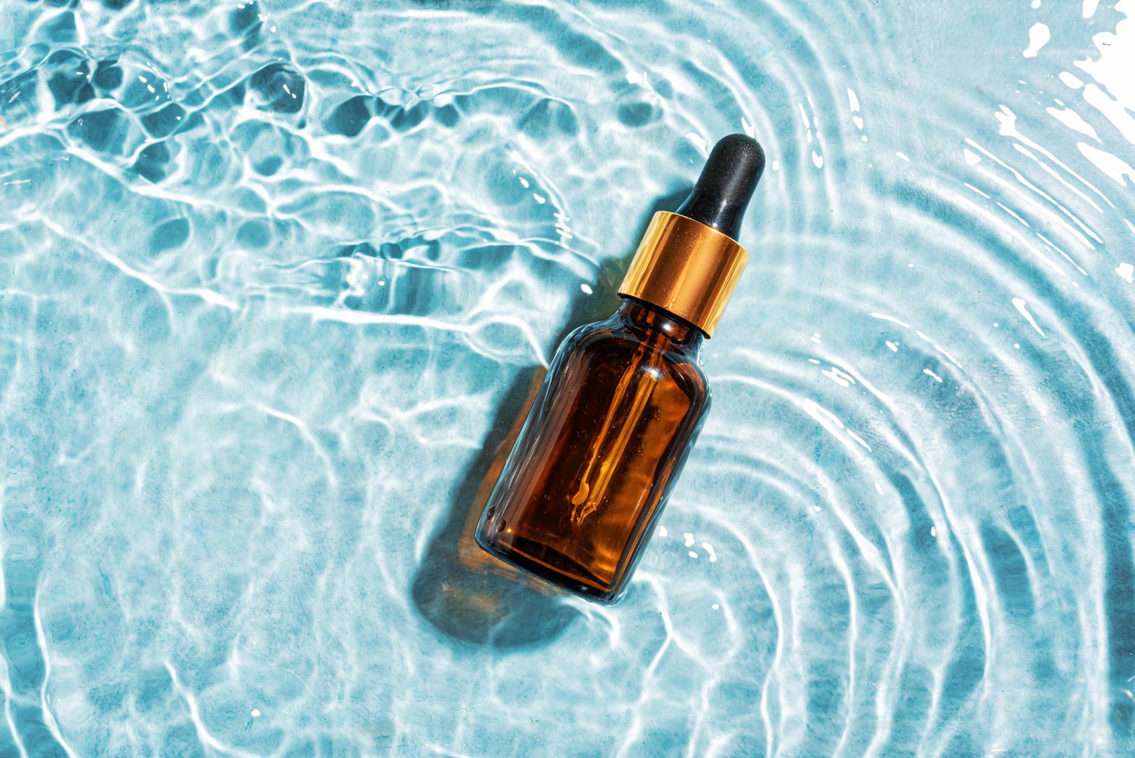 Dropper bottle of cosmetic serum in clear splashing blue water surface, copy space. Moisturizing collagen beauty product, hydrating face treatment.