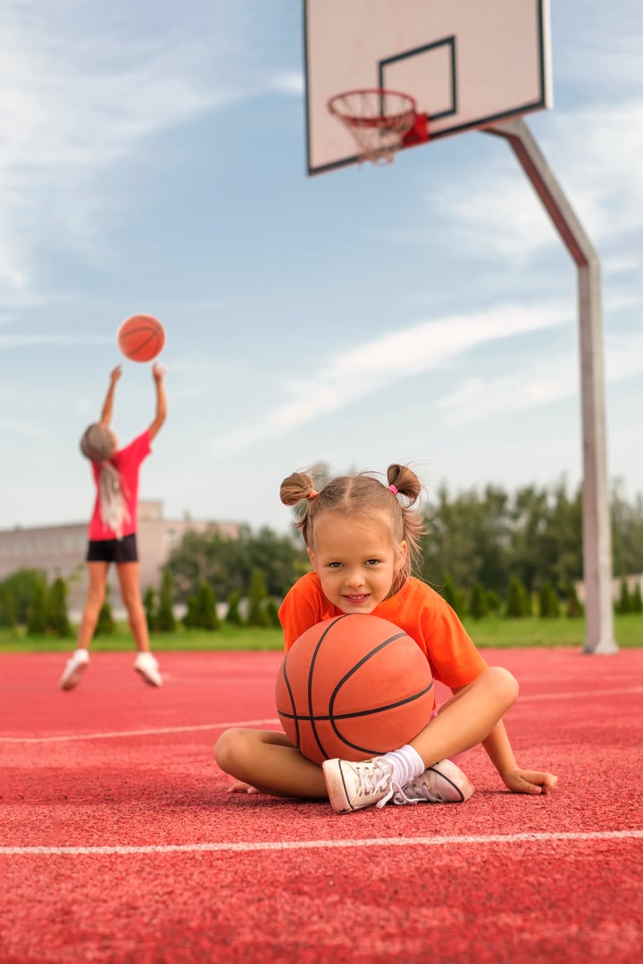 A beautiful cute preschool girl sits with a ball on the playground while her sister throws a ball into a basketball hoop