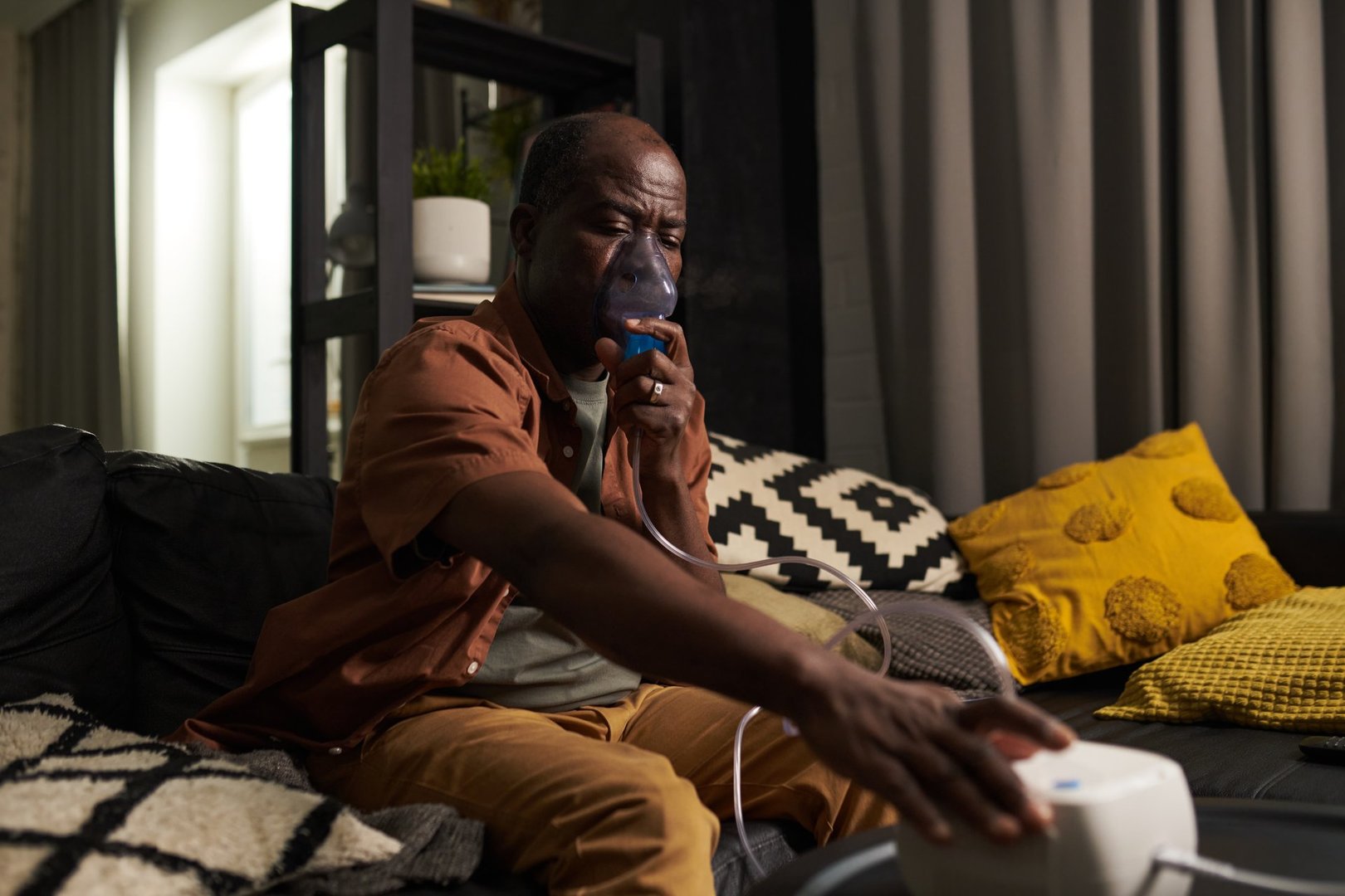 Senior African American man with mask by nose switching on nebulizer and breathing oxygen during medical treatment at home