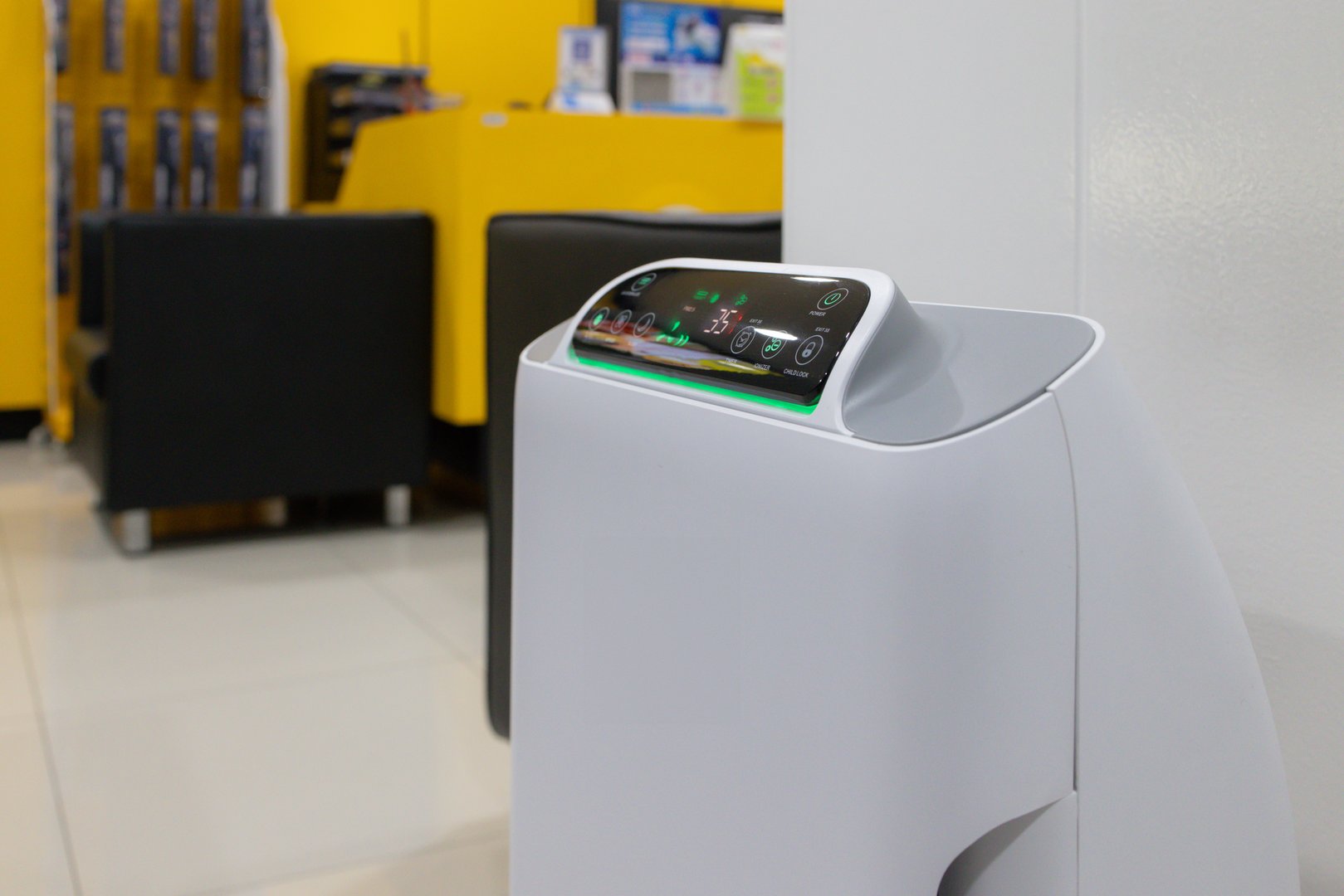 A white device with a green screen sits on a white stand. The device is a small air purifier with a screen that displays the time and a clock. The device is placed in a room with a couch and a chair