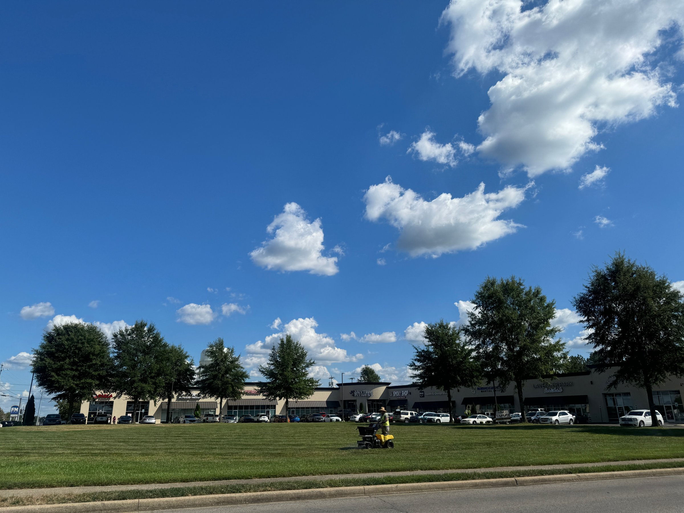 On September 19, 2024, a man mows grass on a lush green lawn in Clarksville, TN, surrounded by trees and a vibrant blue sky filled with fluffy clouds.
