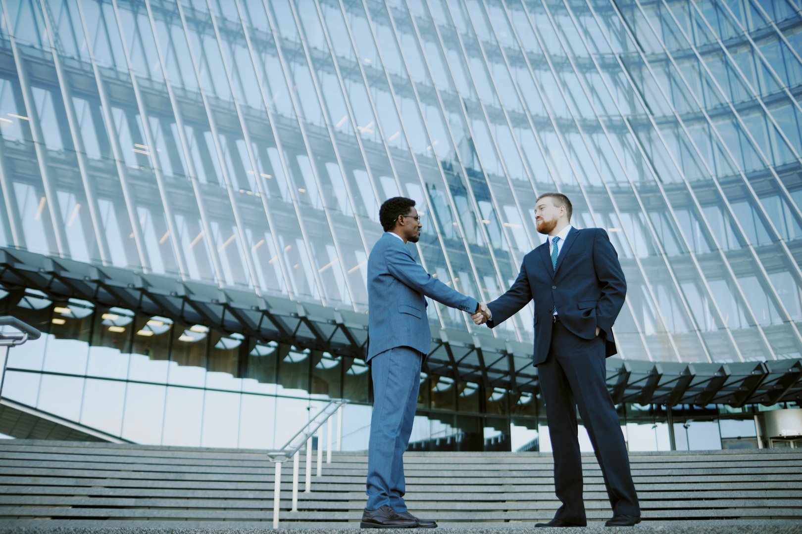 Two businessmen shaking hands while standing outside a modern office building, signifying a professional agreement or partnership