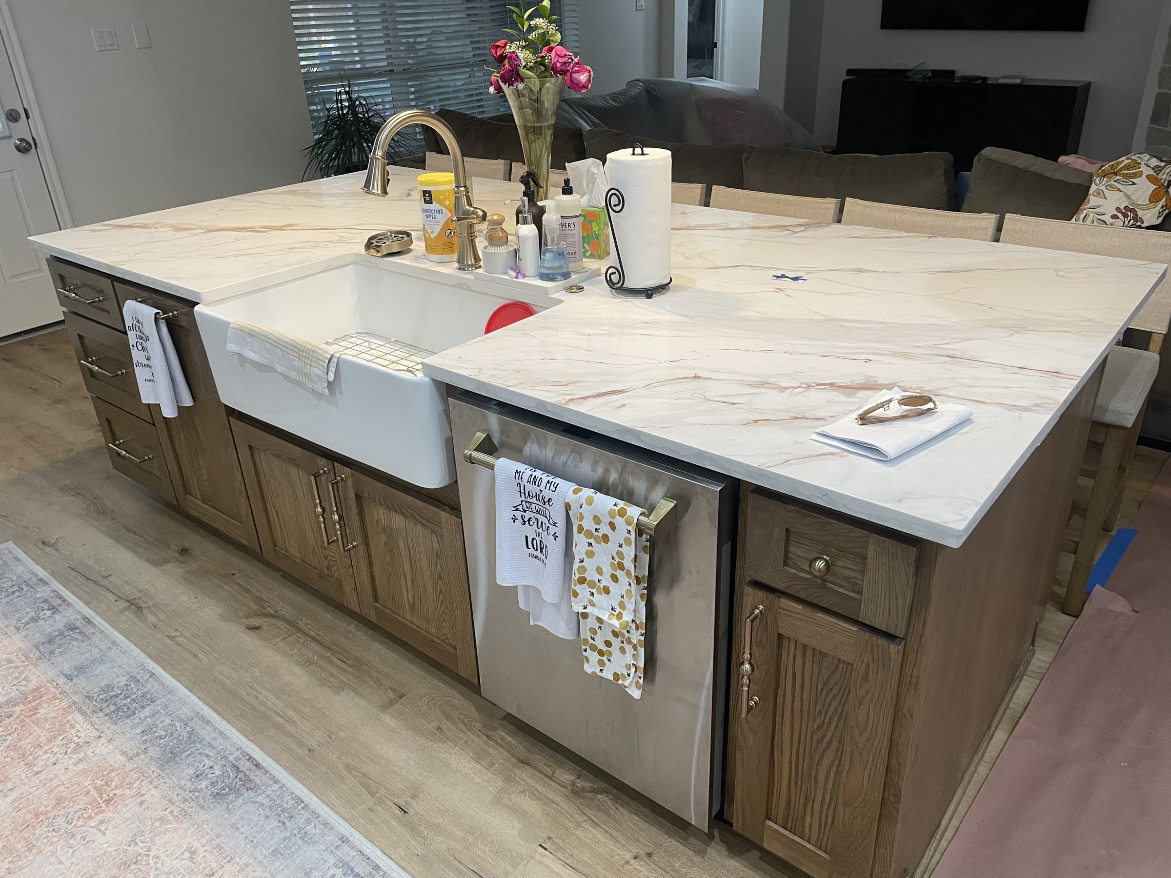 A kitchen island with a marble countertop, farmhouse sink, and various items including flowers, soap, and a paper towel holder.