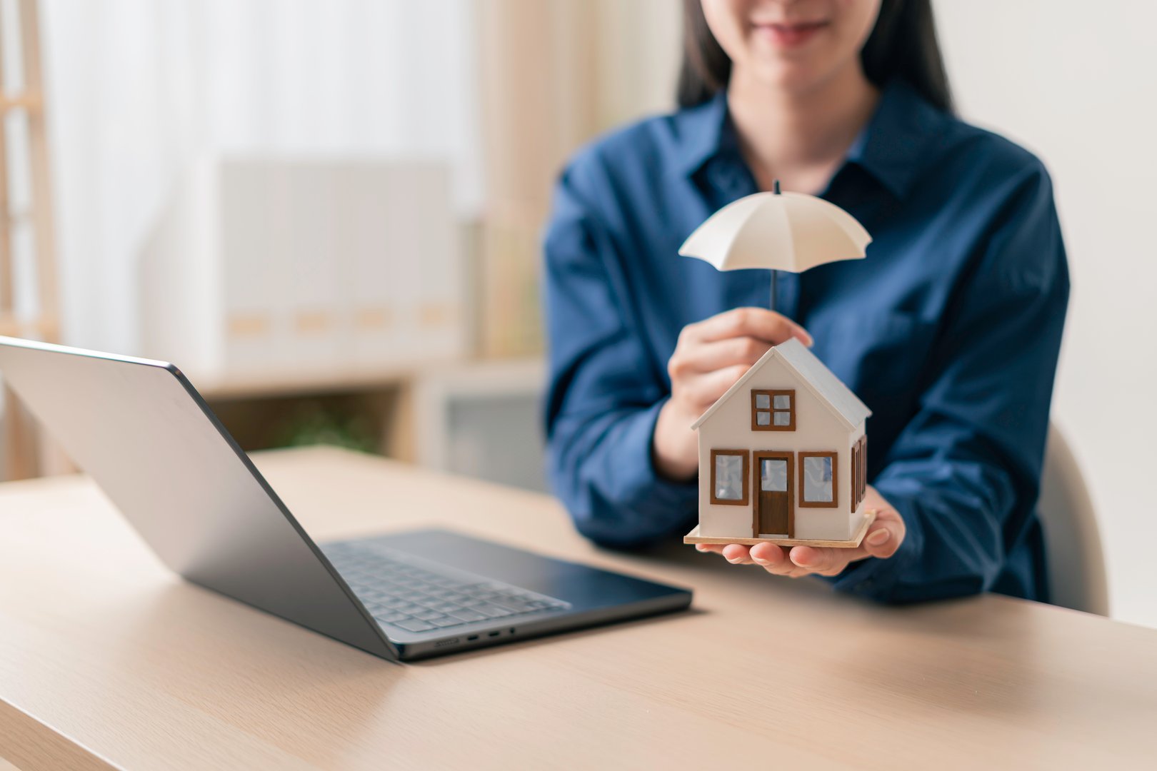 homeowner holding a miniature house under a small umbrella beside a laptop in a home office to illustrate online home insurance and property protection