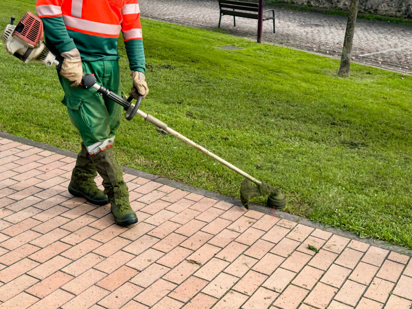 Male worker using trimmer for lawn maintenance in urban park.