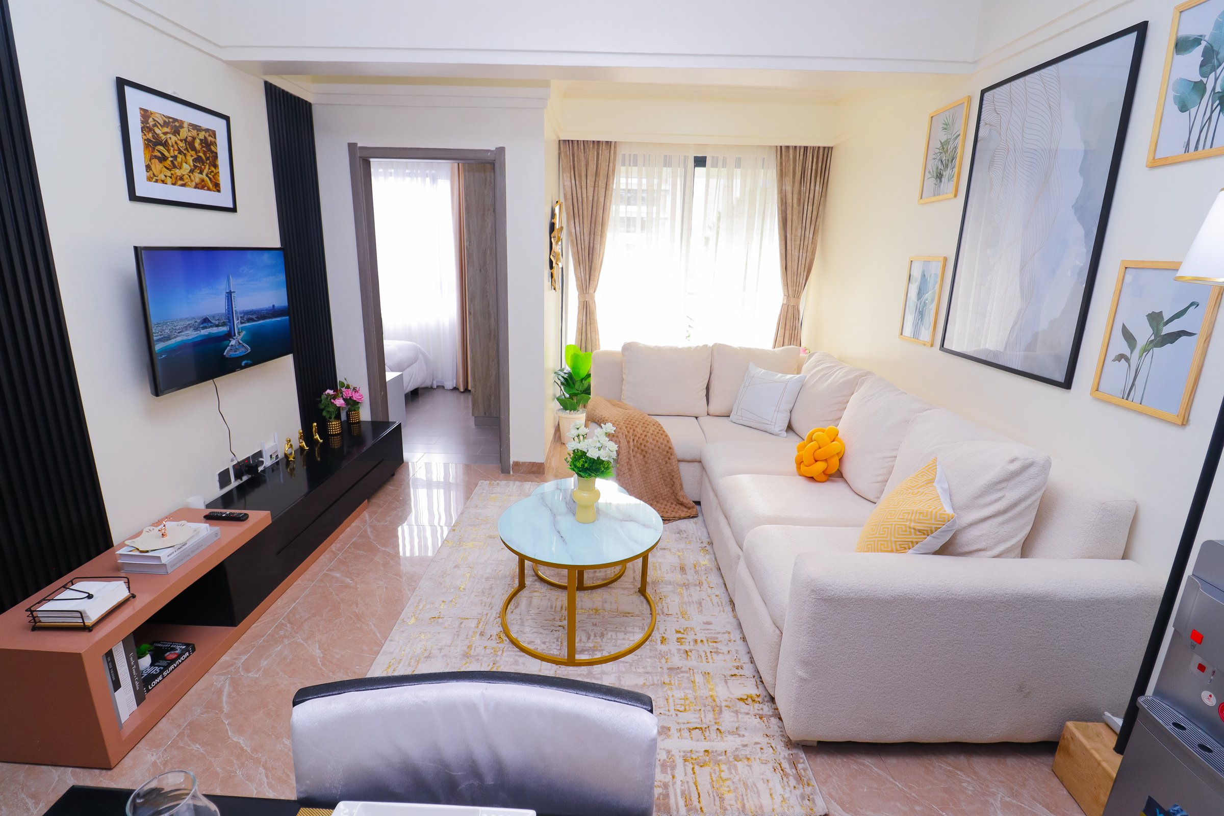 Modern living room with a beige sofa, wall art, coffee table, and TV. Bedroom visible through an open door.