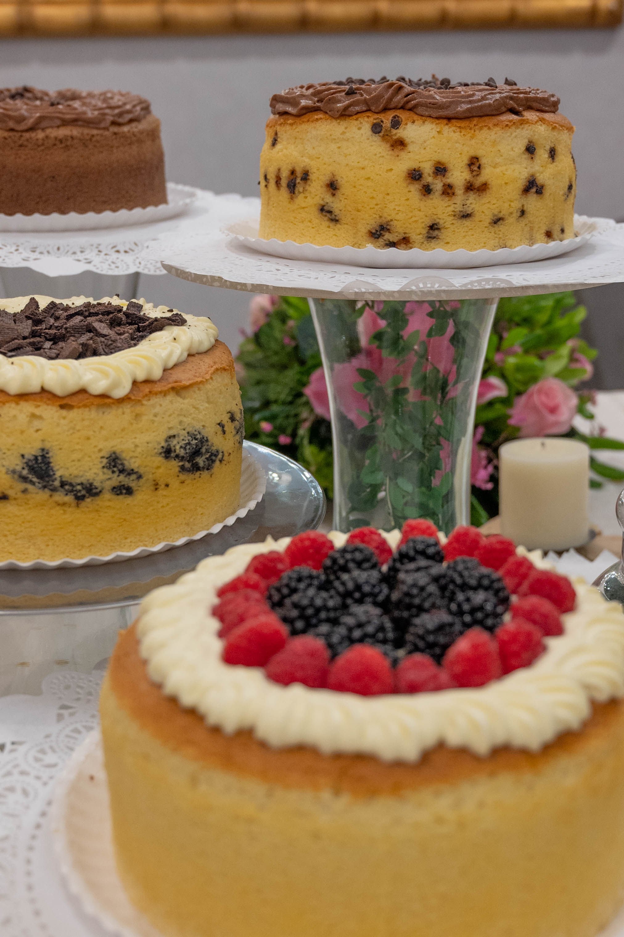 Assorted cakes on stands, featuring vanilla with berries, chocolate chip, and chocolate varieties, with floral decor.
