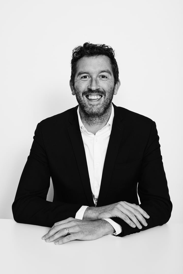 A man in a suit sitting at a table, smiling, with his hands folded. The background is plain white.