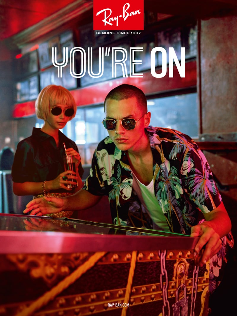 Two people wearing sunglasses indoors, standing by a pinball machine, with a "You're On" Ray-Ban ad theme.