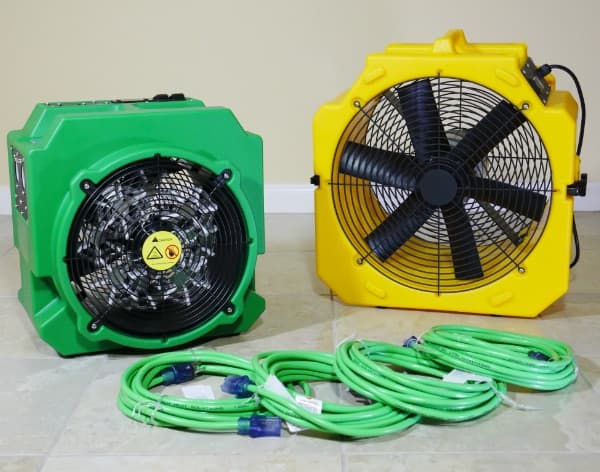 Two industrial fans, one green and one yellow, with electrical cords on a tiled floor against a beige wall.