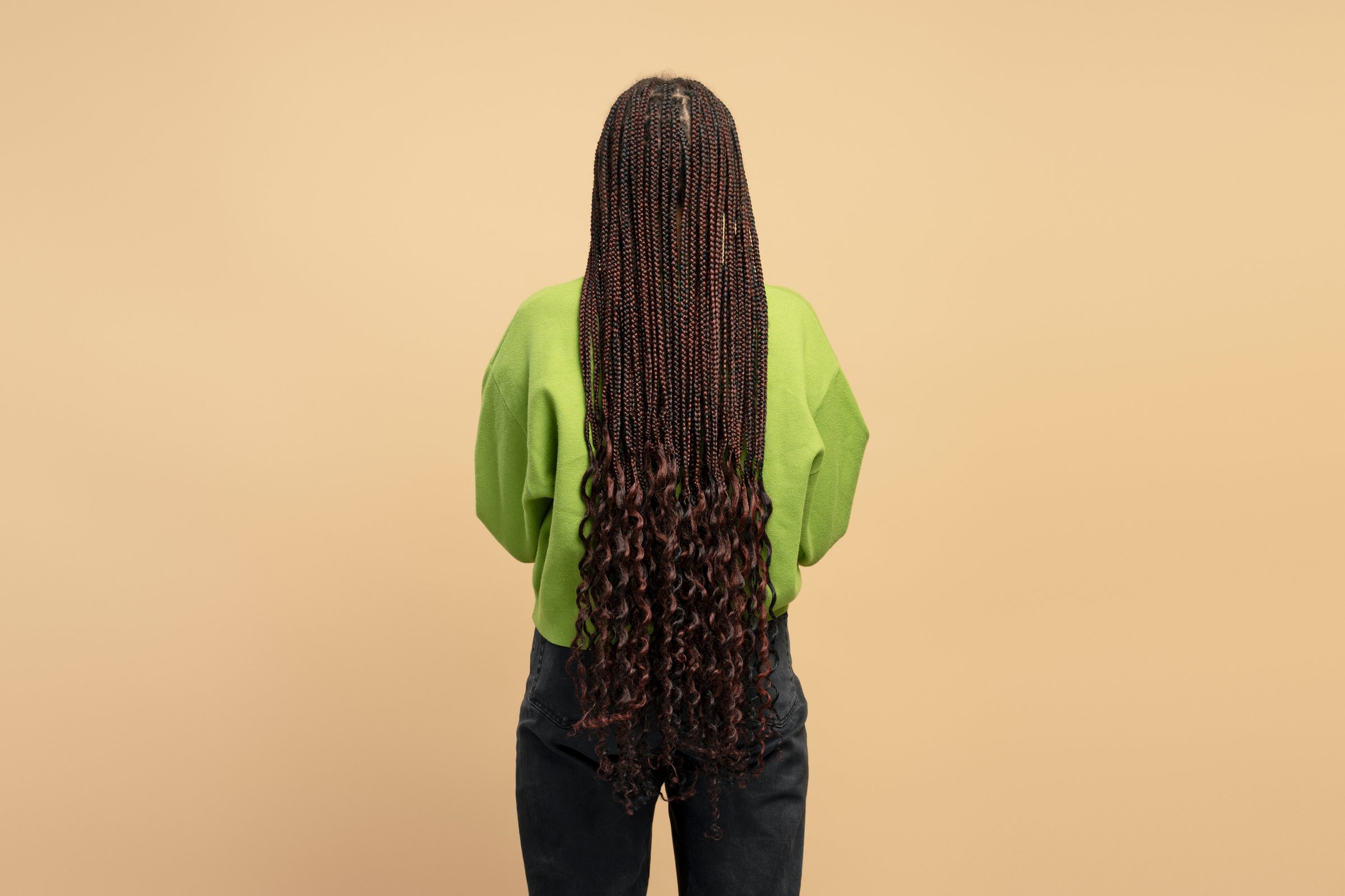 Rear view of a young woman with long braided hair wearing a green sweater and black pants standing against a beige background