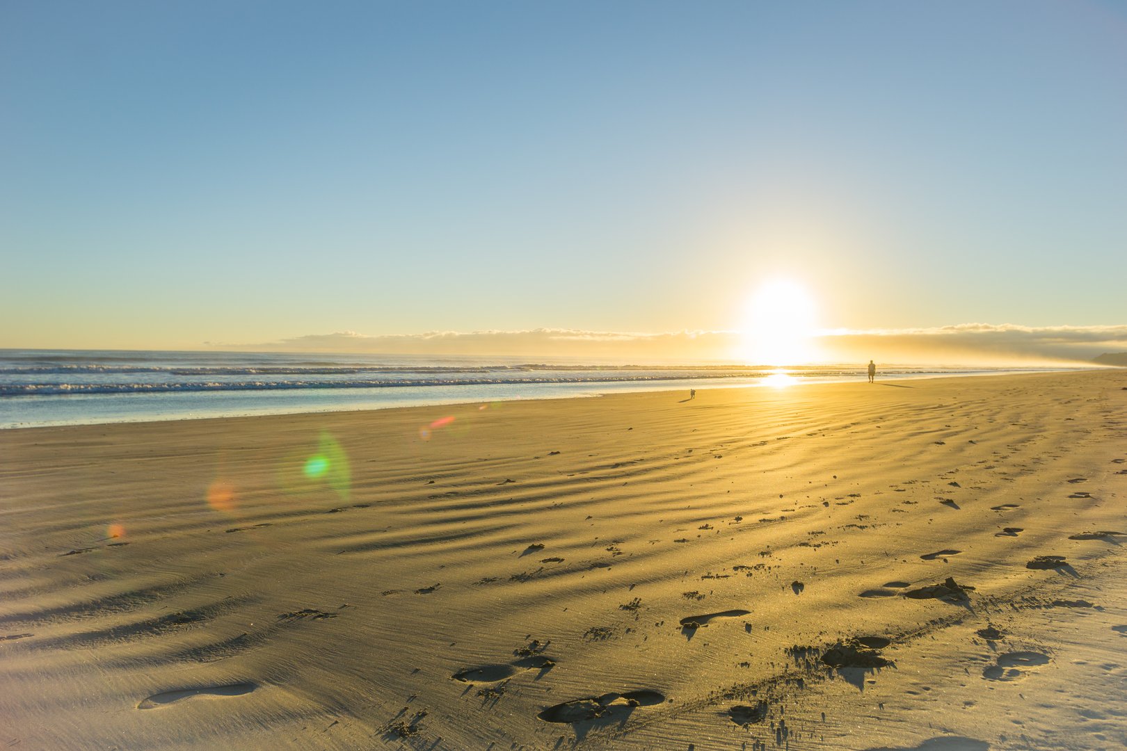 Sunrise over wide flat sandy beach at Ohope Whakatane, New Zealand with distant silhouette of man walking small dog.