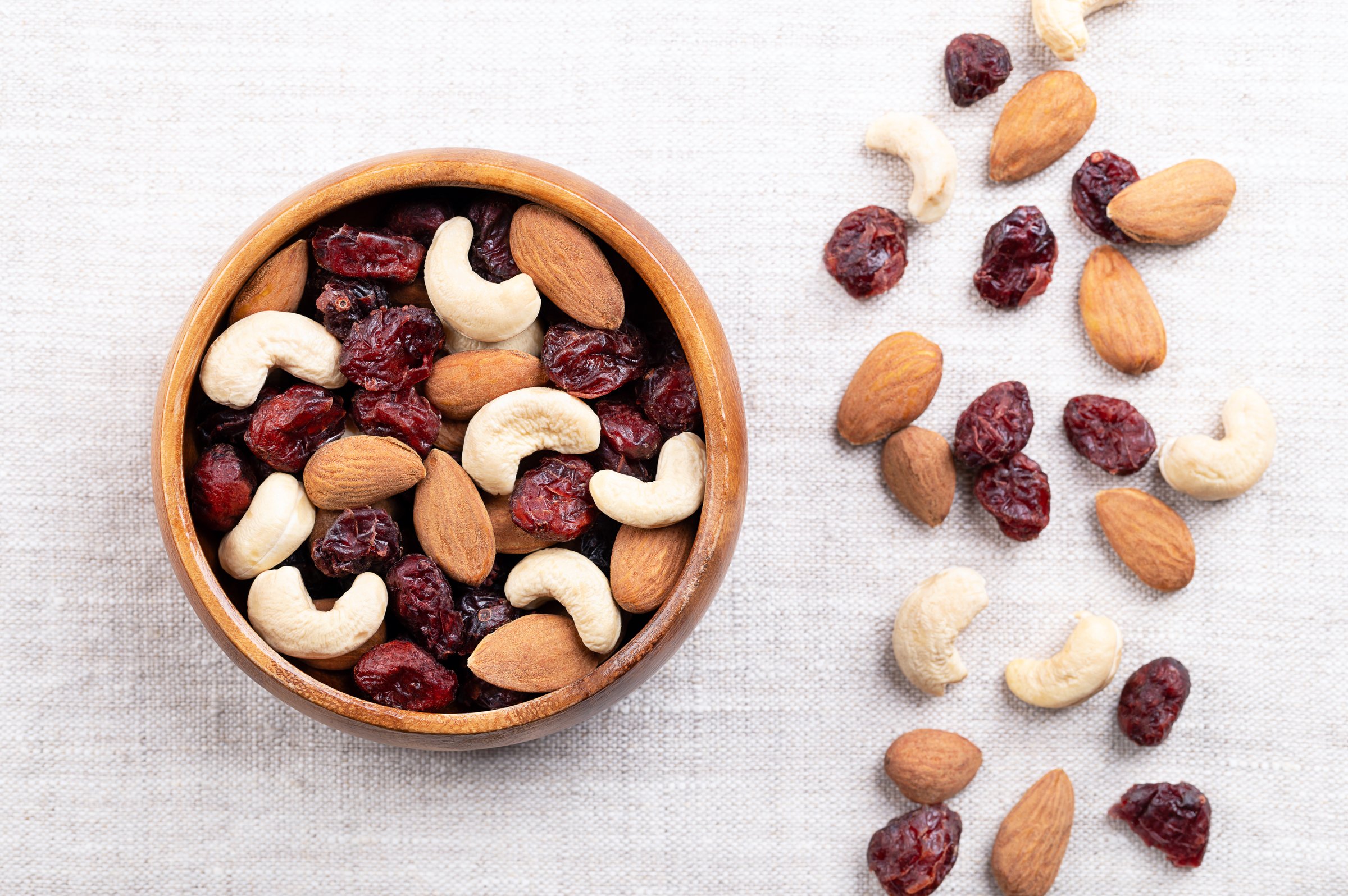 Cranberry almond cashew trail mix in a wooden bowl on linen. Dried sweetened cranberries, with raw almonds, and cashews. Popular snack food, rich on protein, and good source of vitamins and minerals.