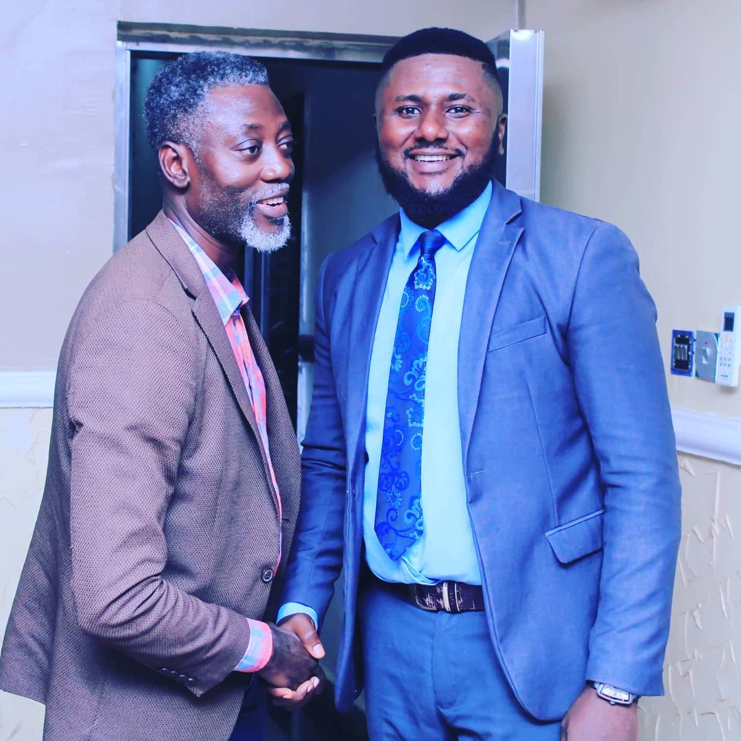 Two men in suits shaking hands and smiling in an indoor setting.