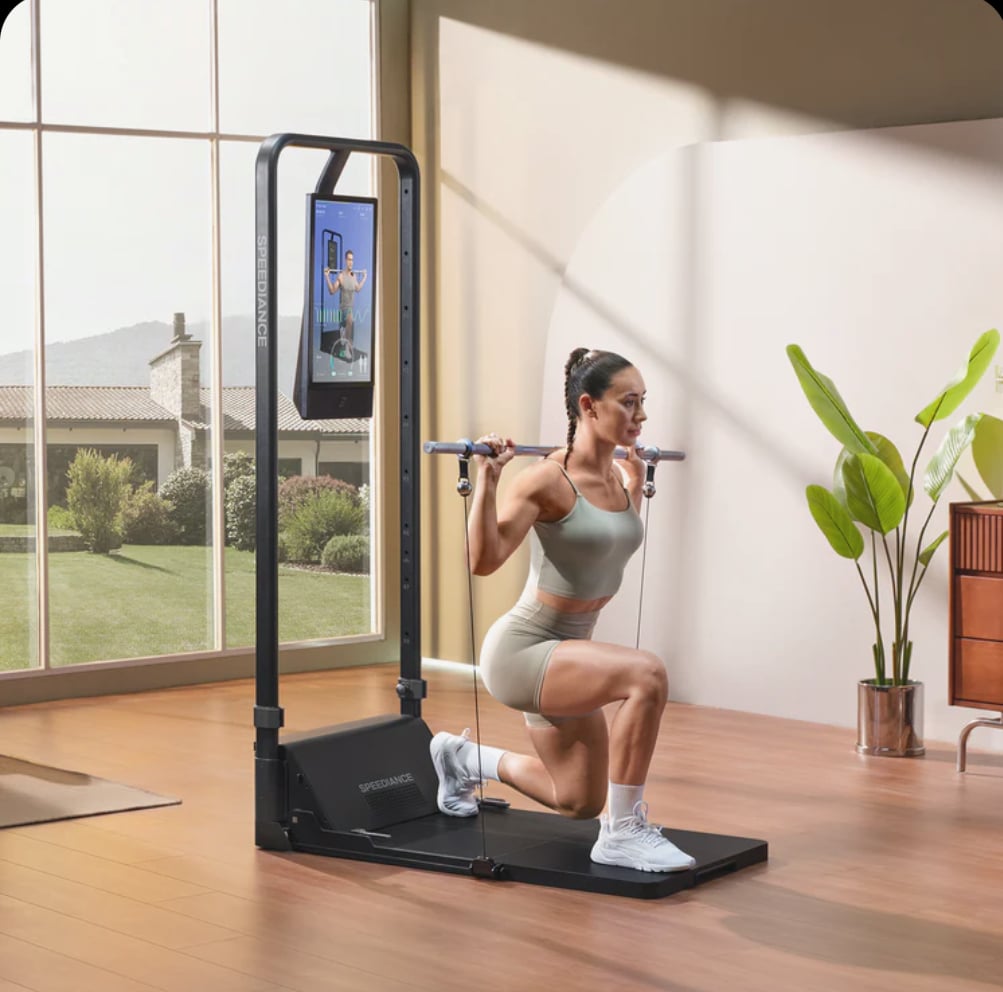 A woman performing a squat with a home fitness machine in a bright living room with large windows, plants, and wooden flooring.