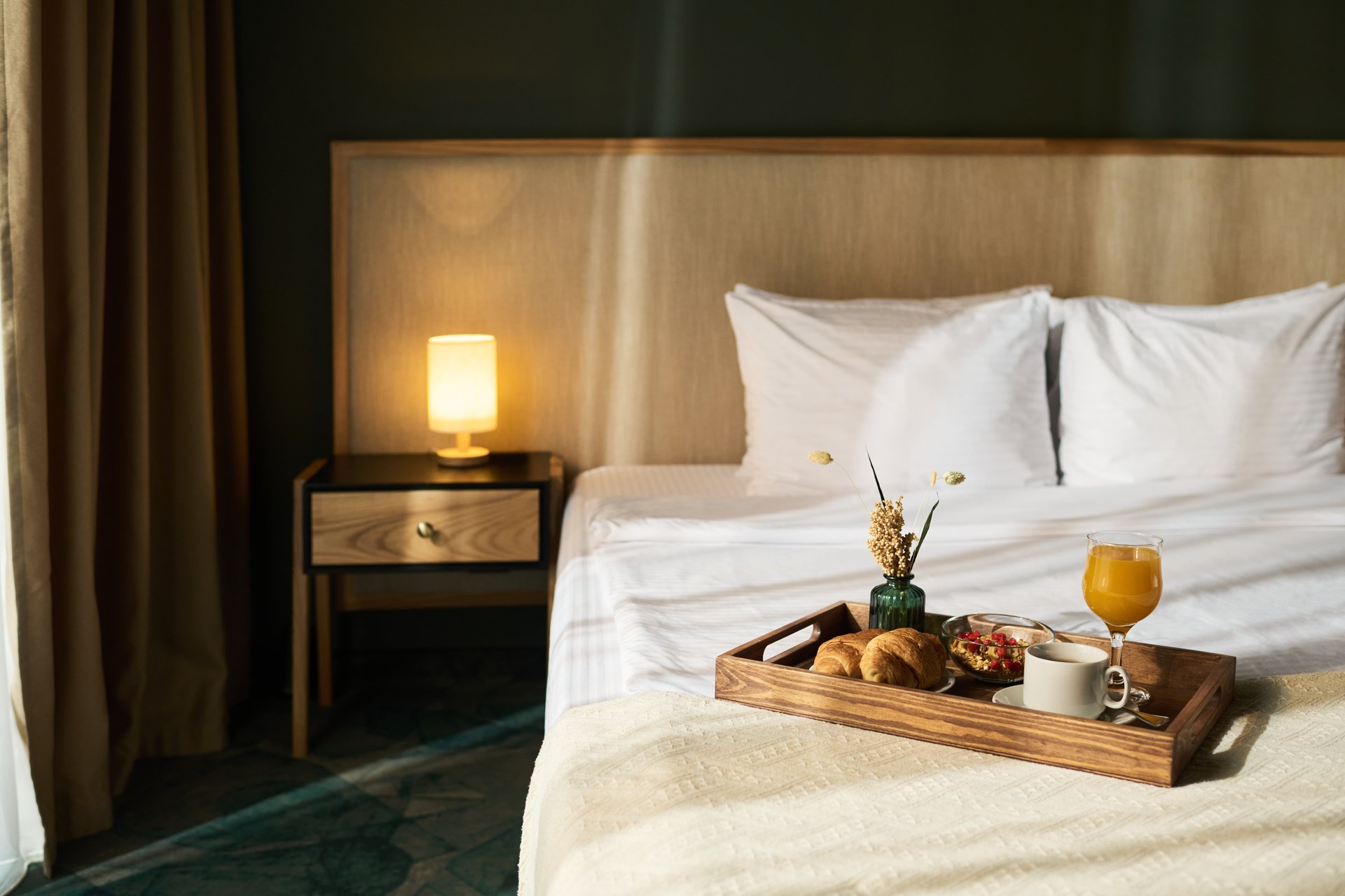 Cozy hotel room showcased with breakfast tray on bed. Tray filled with coffee, juice, pastries, and fruit, with warm lamp providing ambient lighting next to bed