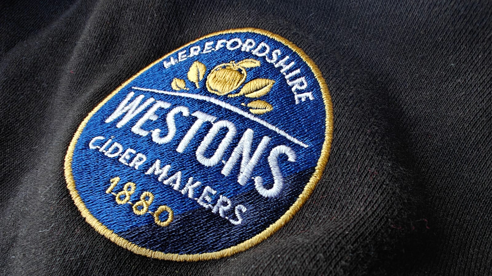 Embroidered patch reading Westons Cider Makers 1880 on a black fabric, featuring a gold apple emblem.