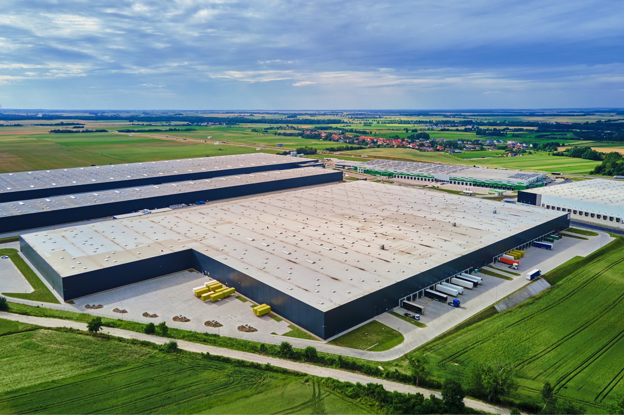 Large modern warehouse with loading docks and parked trucks surrounded by green fields. Industrial logistics hub. Transport infrastructure for distribution, shipping and storage in supply chain system