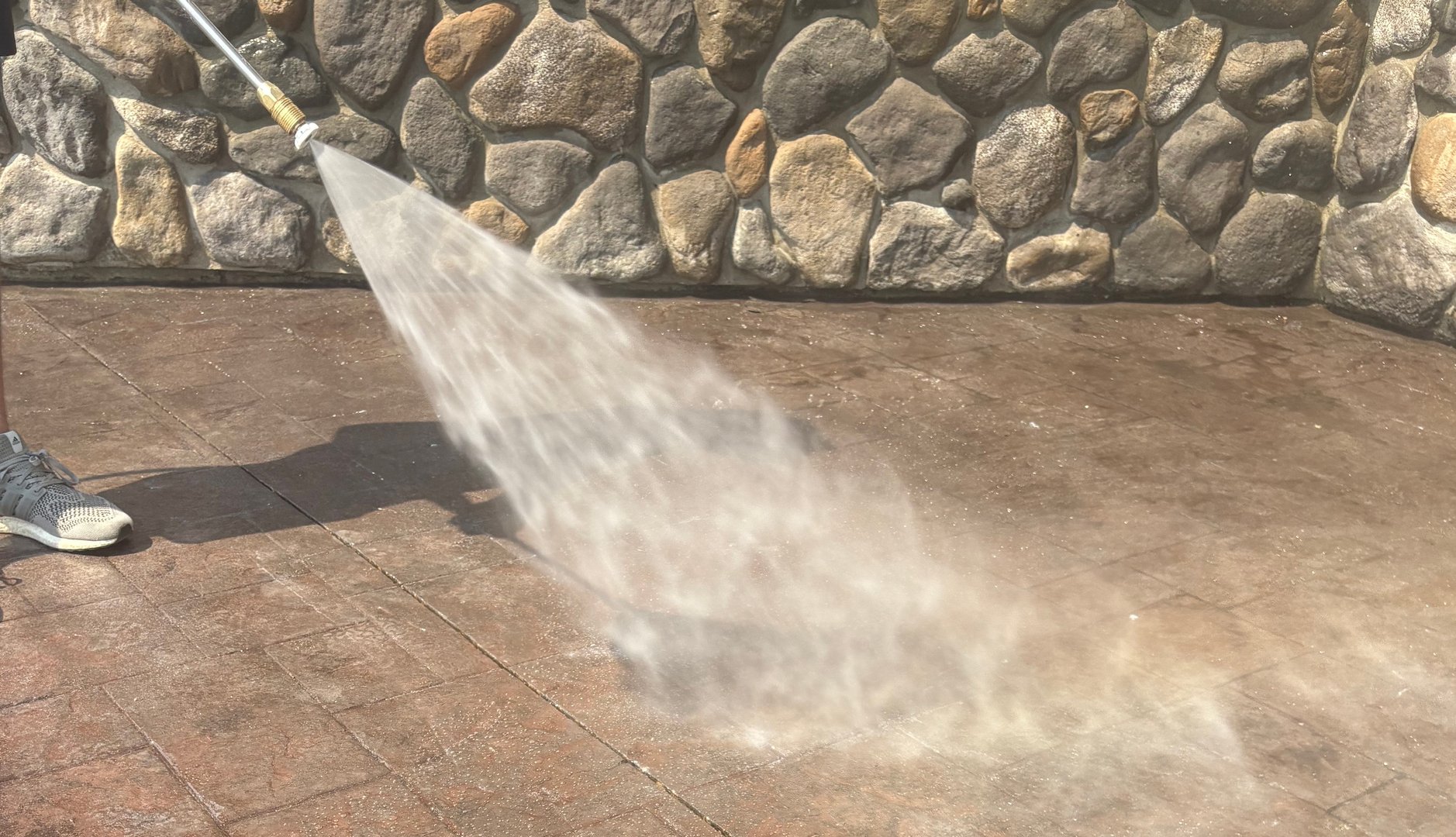 Person pressure washing a stone patio with a spray nozzle, removing dirt and grime, stone wall in the background.