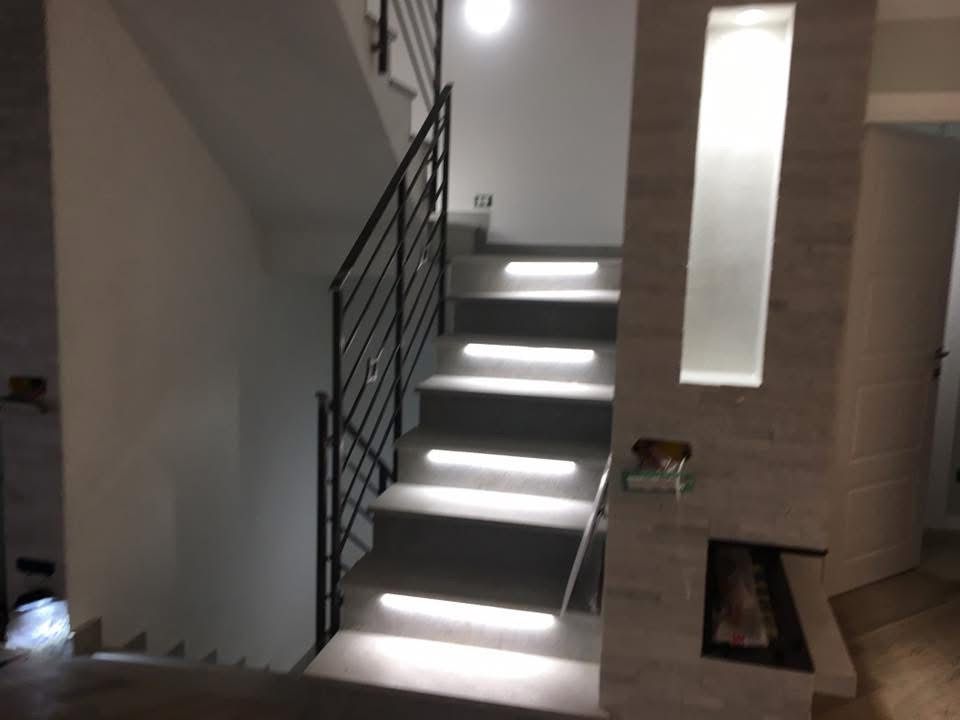 Modern staircase with illuminated steps and a black metal railing, positioned next to a stone-textured wall.