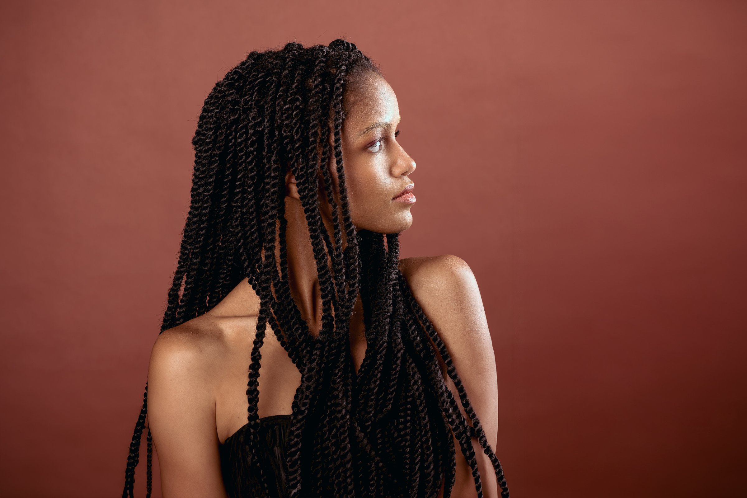 Portrait of thoughtful young African American female with long Afro braids and bare shoulders looking away while sitting in room against brown background in light