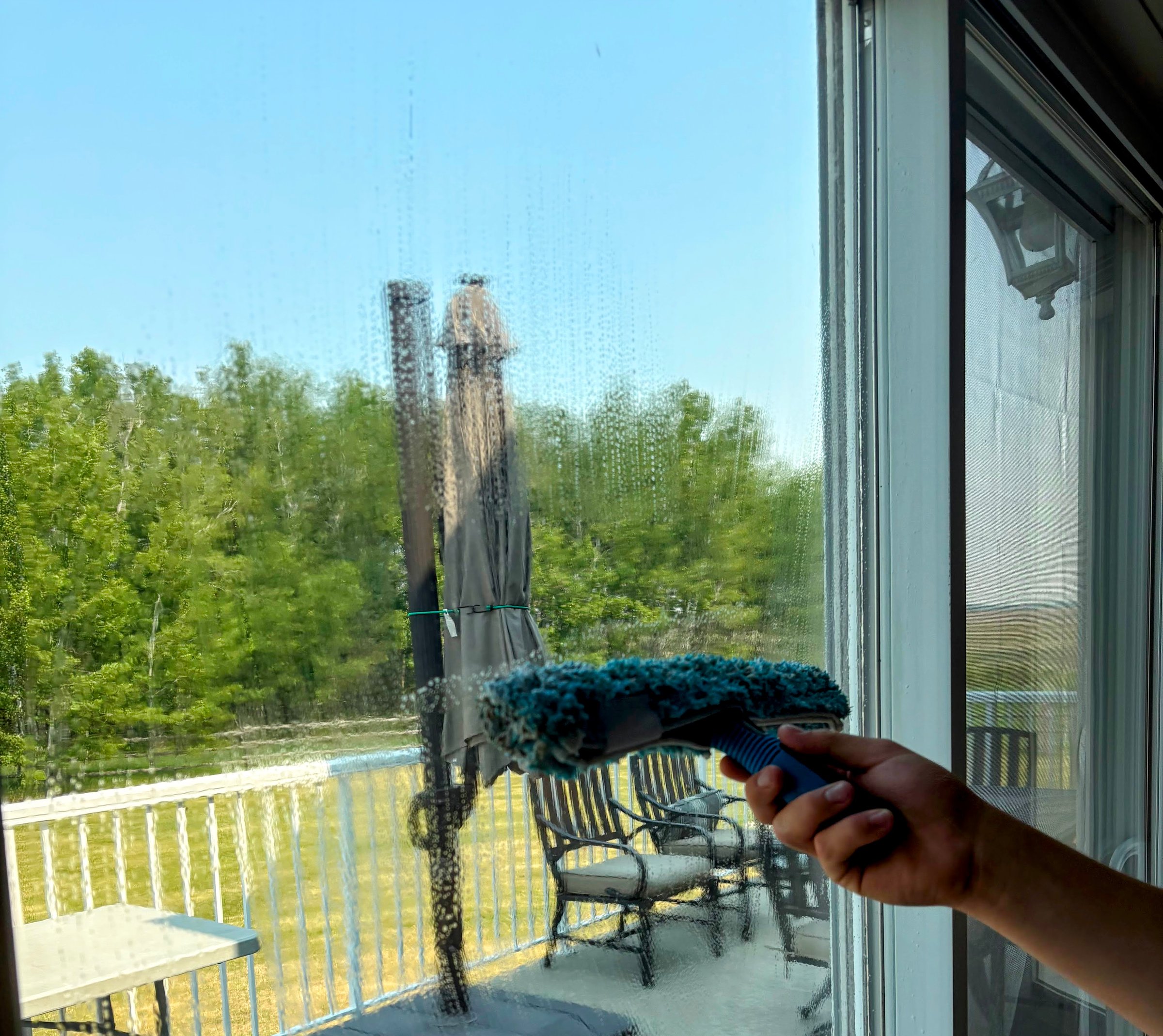 Person cleaning a glass door with a squeegee, outdoor view of patio furniture and trees in the background.