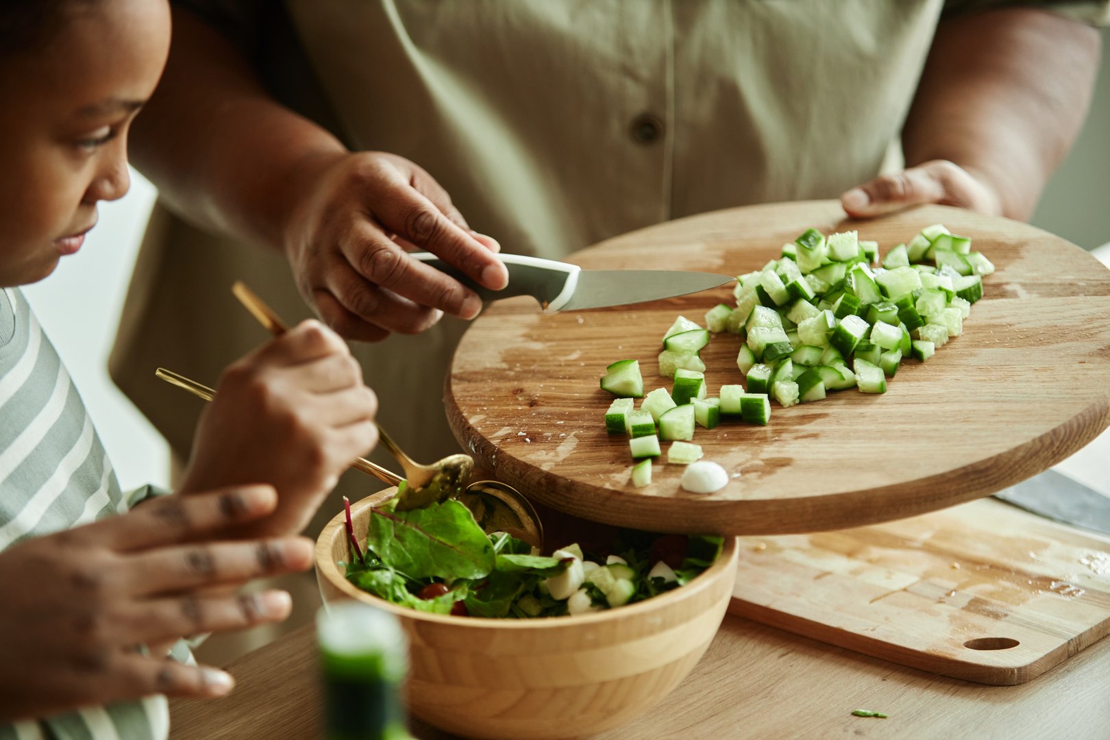 Close up on hands of Black senior woman adding chopped cucumbers to green lettuce leaves in salad bowl using wooden cutting board and knife while cooking together with girl at kitchen, copy space