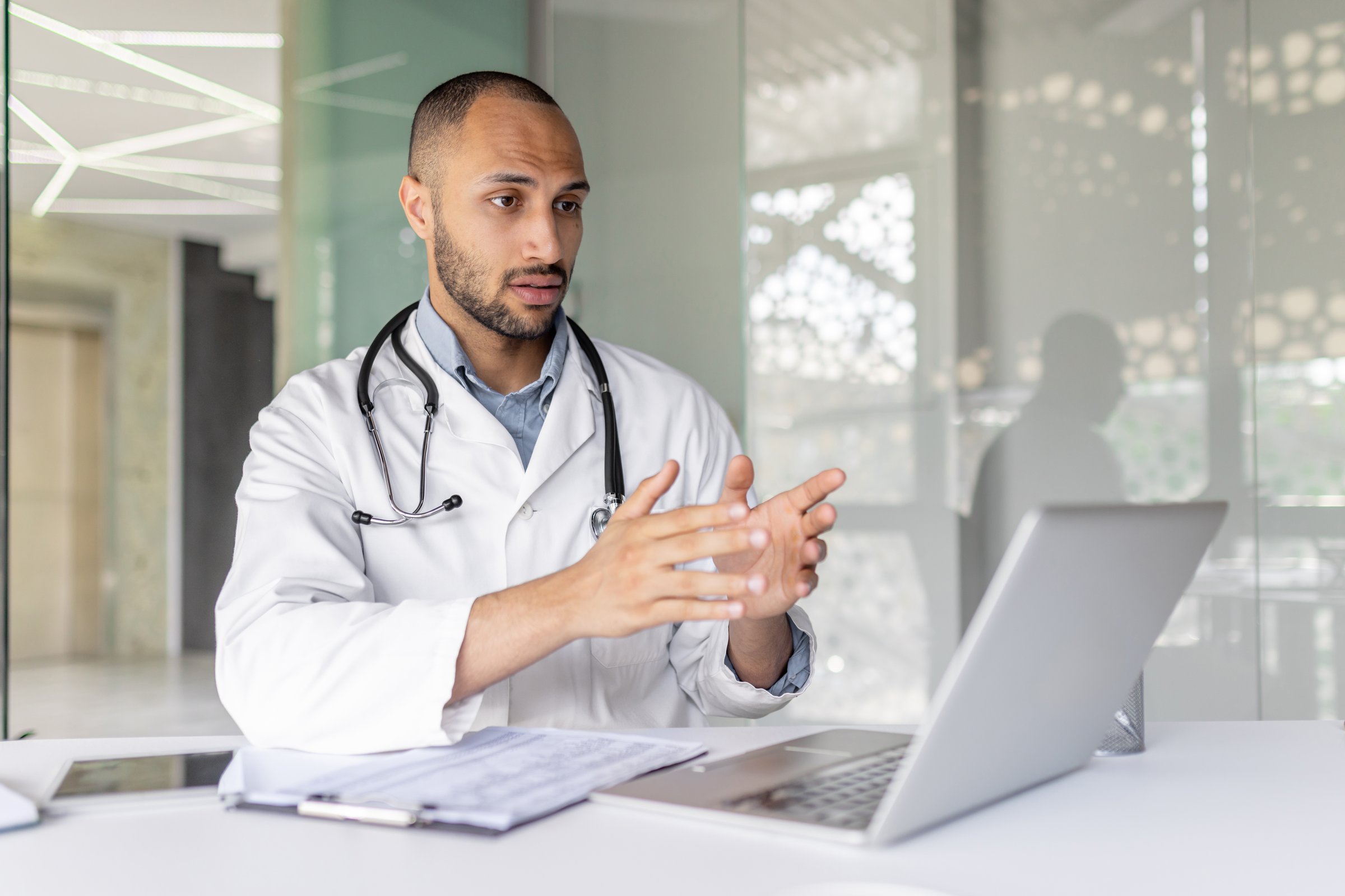 A doctor, wearing a lab coat and stethoscope, gestures while on a video call using a laptop in a modern office setting.