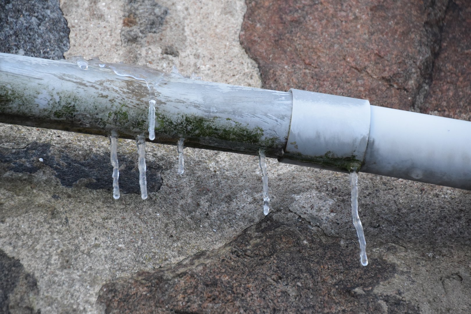 A frozen pipe with hanging icicles against a granite stone wall.