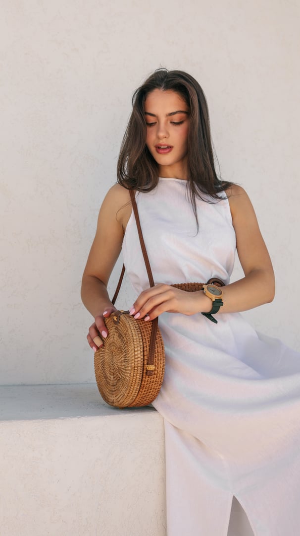 A young woman in a white dress is holding a round rattan bag. She is wearing a watch and a belt, and the background is a white wall.