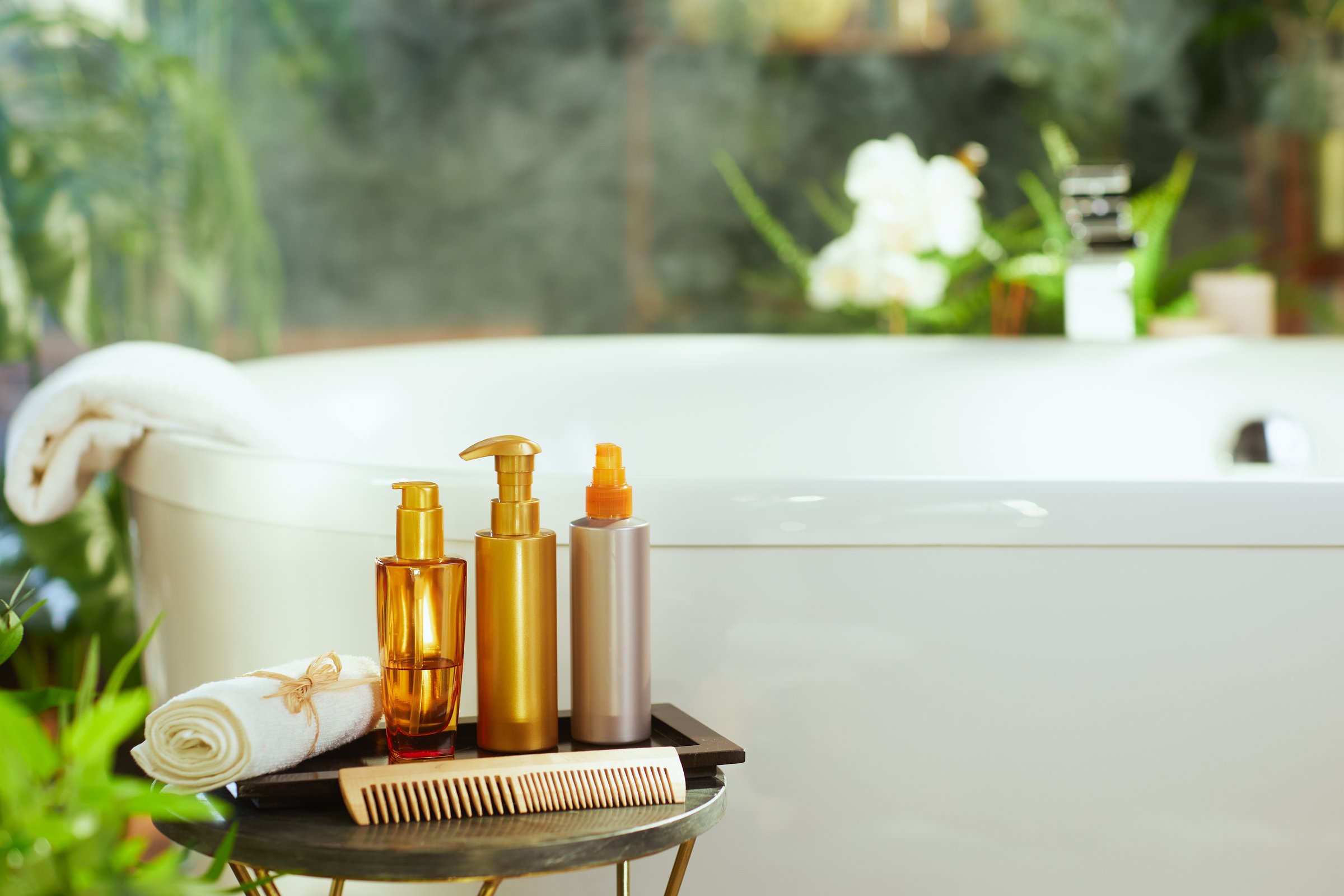 Hair & bath products, bottles, rolled towel, & wooden comb, arranged on a side table. White bathtub & green plants in background convey a tranquil, luxurious self-care experience.