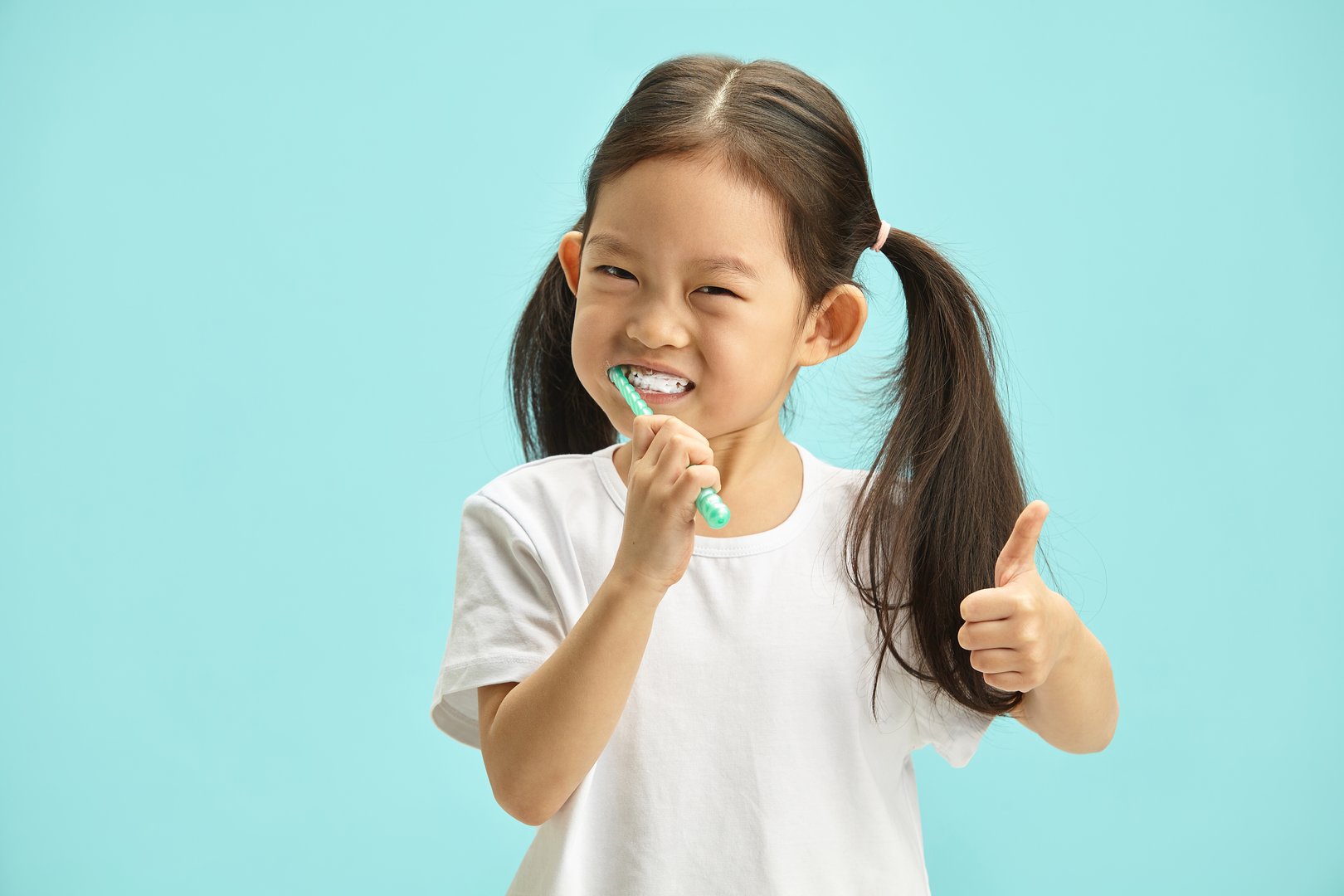 Cute Chinese little girl having perfect smile brushing her teeth with plastic toothbrush on blue background, keep thumb raised, studio portrait. Children health and oral care concept.