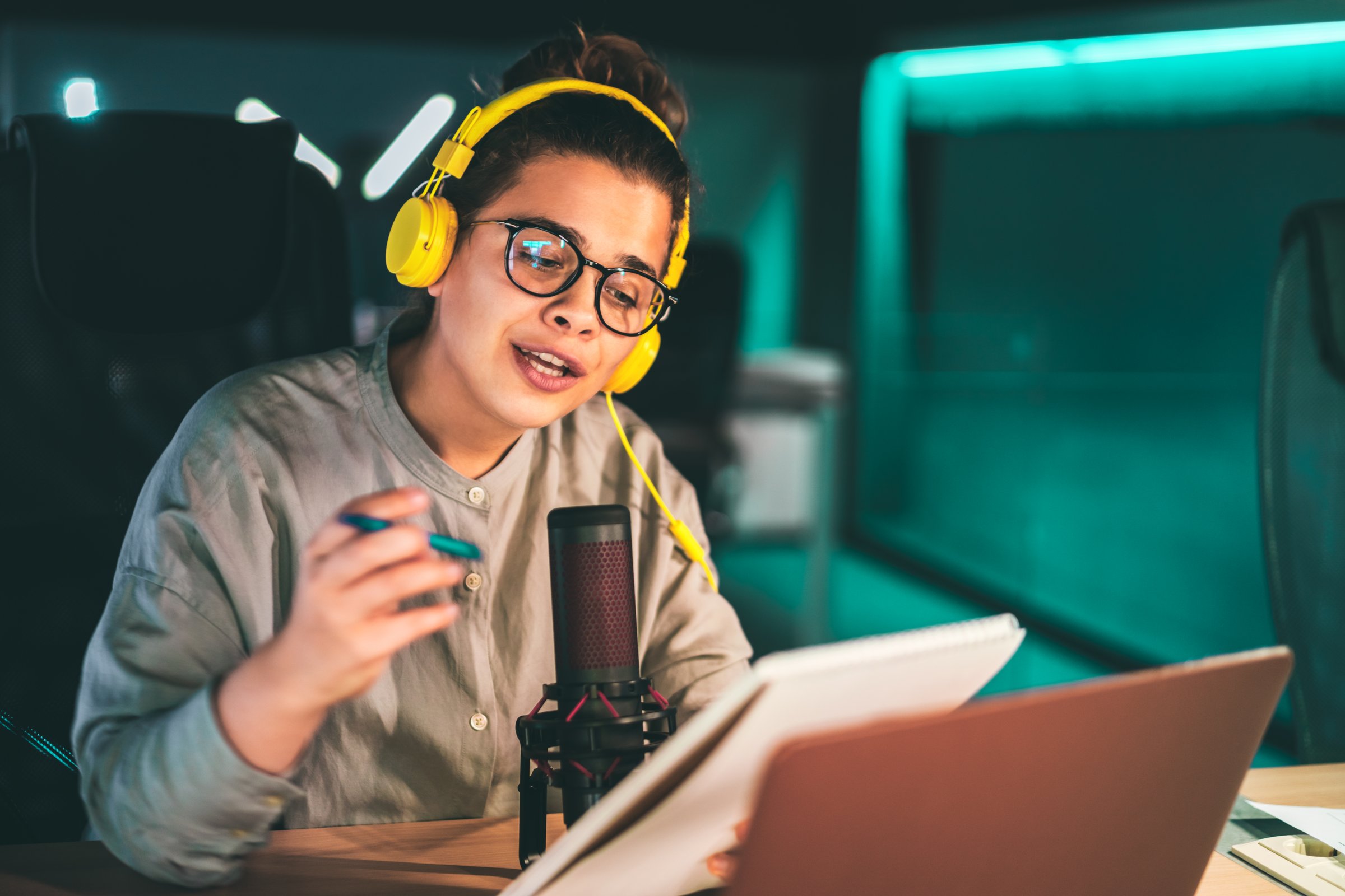 Young woman recording an audio podcast in a modern studio, wearing headphones and hipster glasses, speaking to the microphone and holding a notepad